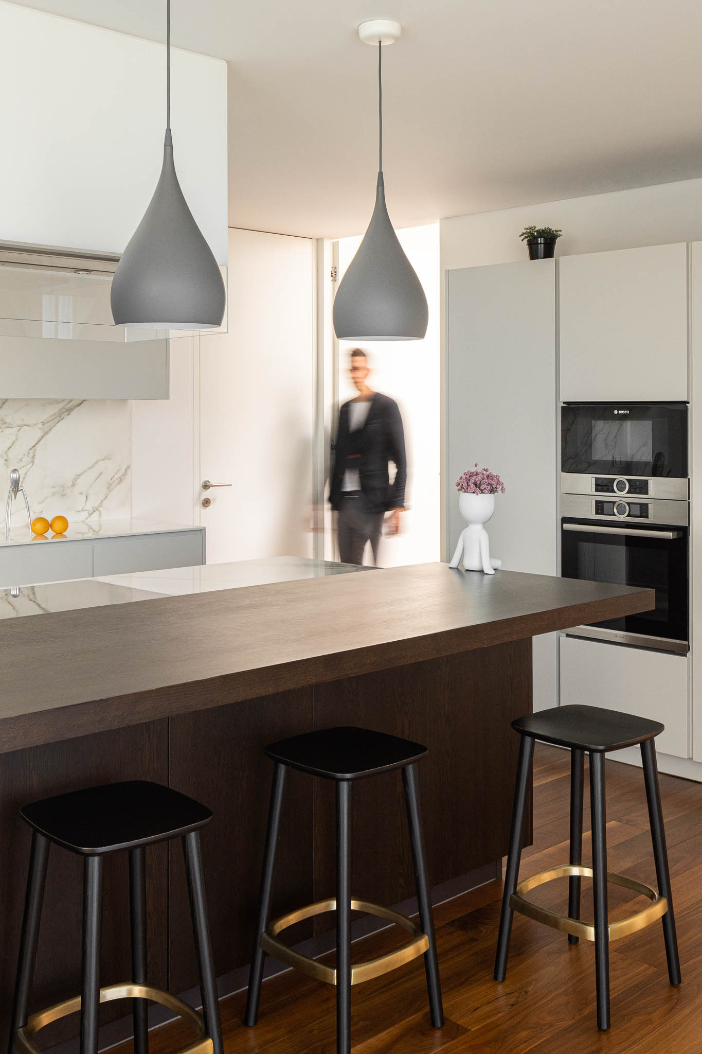 Cozinha moderna com ilha em madeira escura, bancos pretos com detalhes dourados e candeeiros suspensos cinzentos. — Casa PS arquitetura Inception Architects Studio em Braga