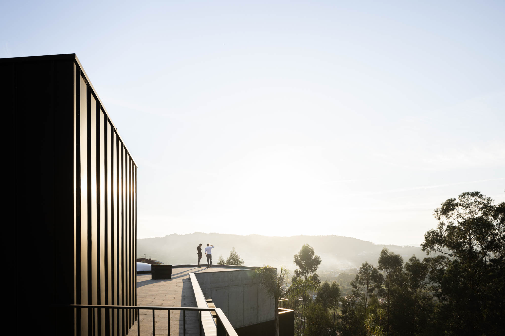 Vista exterior da casa com revestimento em zinco preto e pessoas no terraço, com paisagem montanhosa ao fundo. — Casa PS arquitetura Inception Architects Studio em Braga