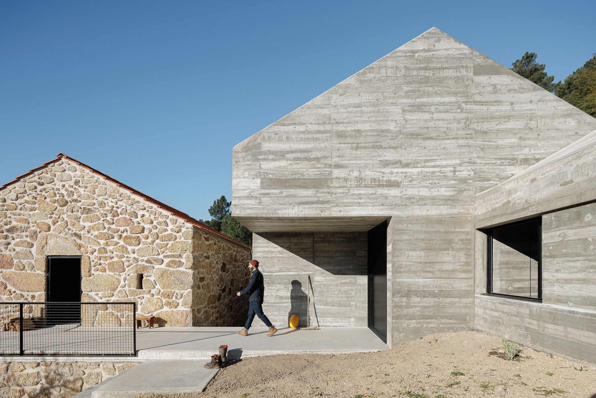 Casa NaMora: Edifício em betão com telhado inclinado, entrada marcada, ao lado de construção rústica em granito. — Casa NaMora arquitetura David Bilo Arquitectos e Filipe Pina Arquitectura em Guarda
