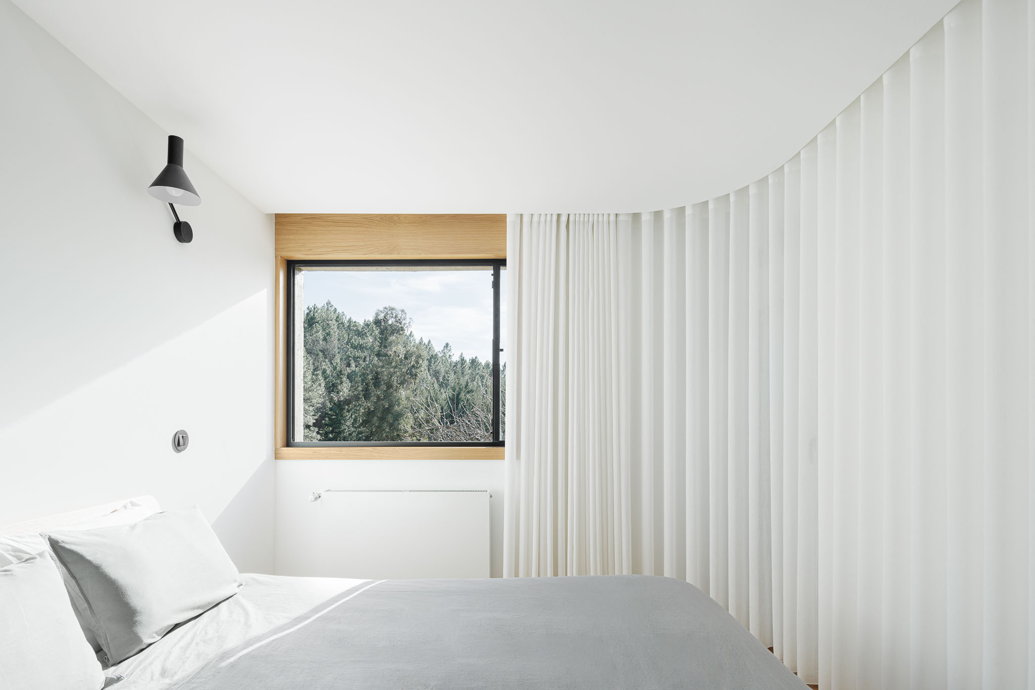 Casa NaMora: Quarto minimalista com cama, cortinas brancas onduladas e janela com vista para a natureza. — Casa NaMora arquitetura David Bilo Arquitectos e Filipe Pina Arquitectura em Guarda