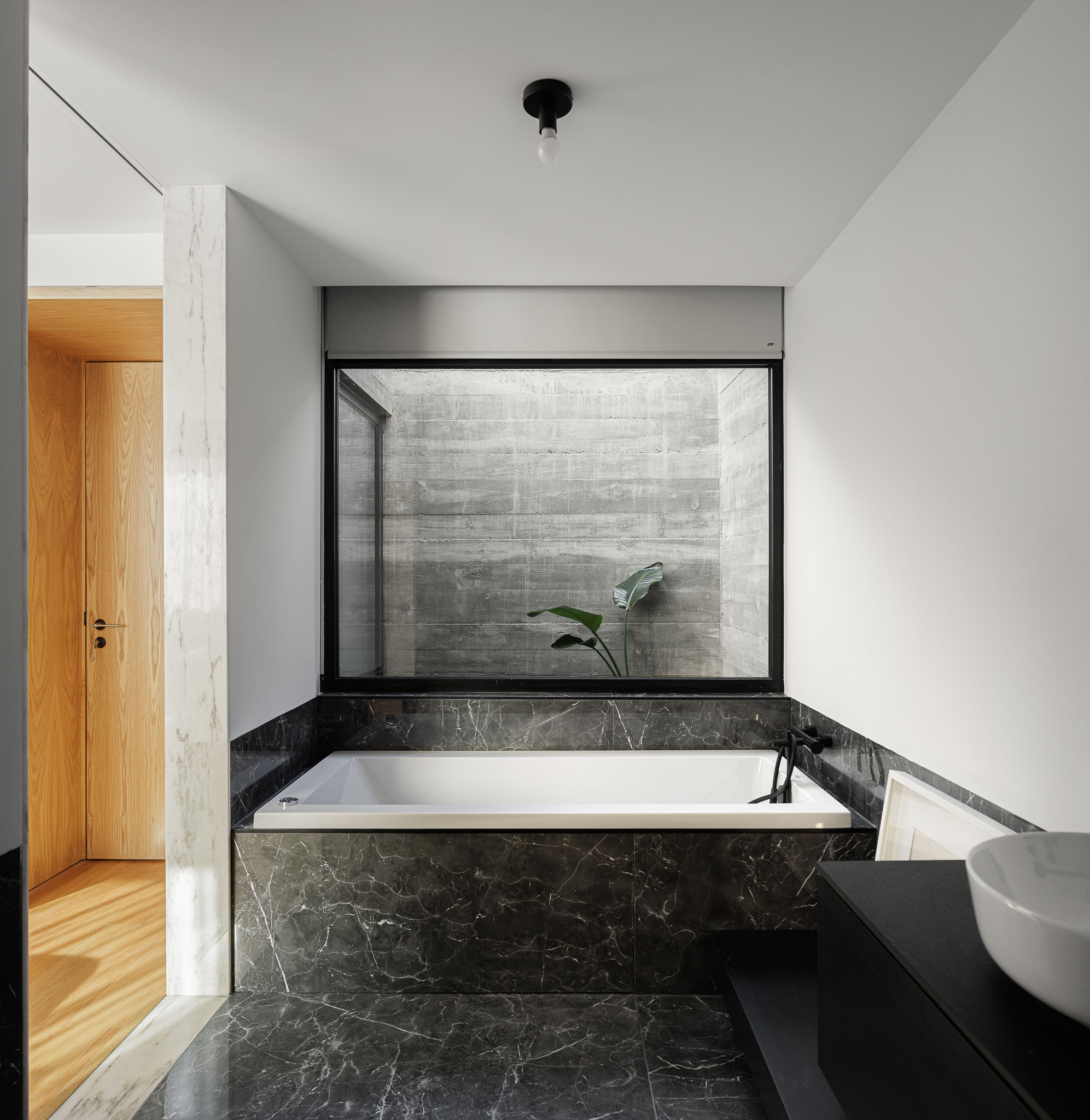 Casa NaMora: Casa de banho com banheira em mármore preto, janela para pátio interior e detalhes em madeira clara. — Casa NaMora arquitetura David Bilo Arquitectos e Filipe Pina Arquitectura em Guarda