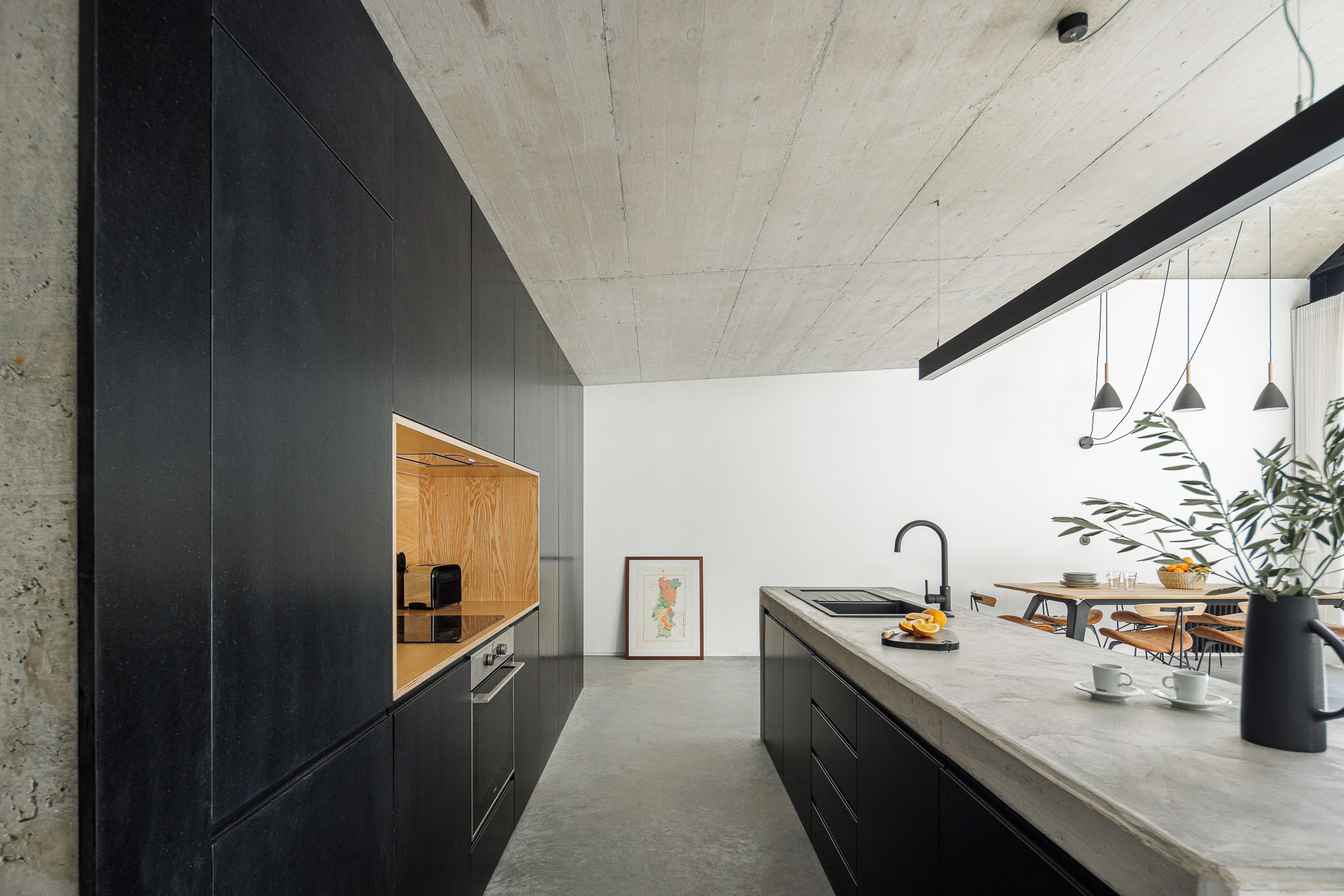 Casa NaMora: Cozinha moderna com armários pretos, bancada em cimento e detalhes em madeira clara, sob teto em betão. — Casa NaMora arquitetura David Bilo Arquitectos e Filipe Pina Arquitectura em Guarda
