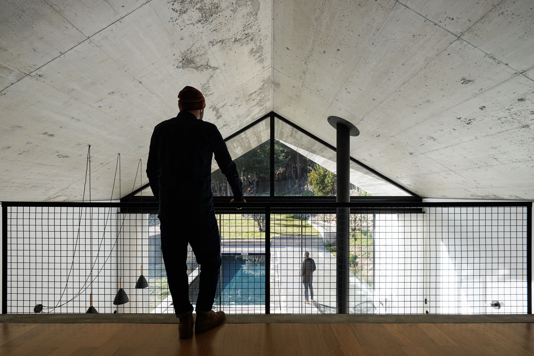 Casa NaMora: Homem no mezanino a observar a sala de estar com lareira e a piscina através da janela envidraçada. — Casa NaMora arquitetura David Bilo Arquitectos e Filipe Pina Arquitectura em Guarda