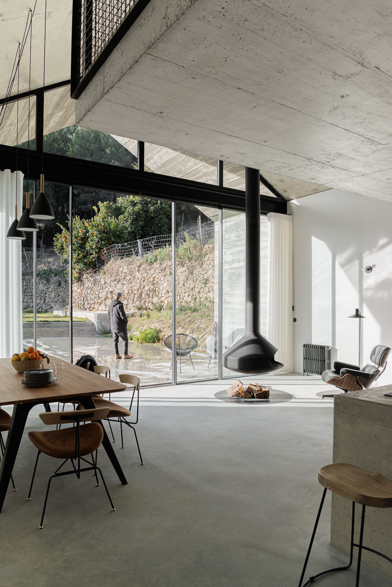 Casa NaMora: Interior da sala com lareira suspensa, mesa de jantar e vista para o exterior através da ampla janela. — Casa NaMora arquitetura David Bilo Arquitectos e Filipe Pina Arquitectura em Guarda