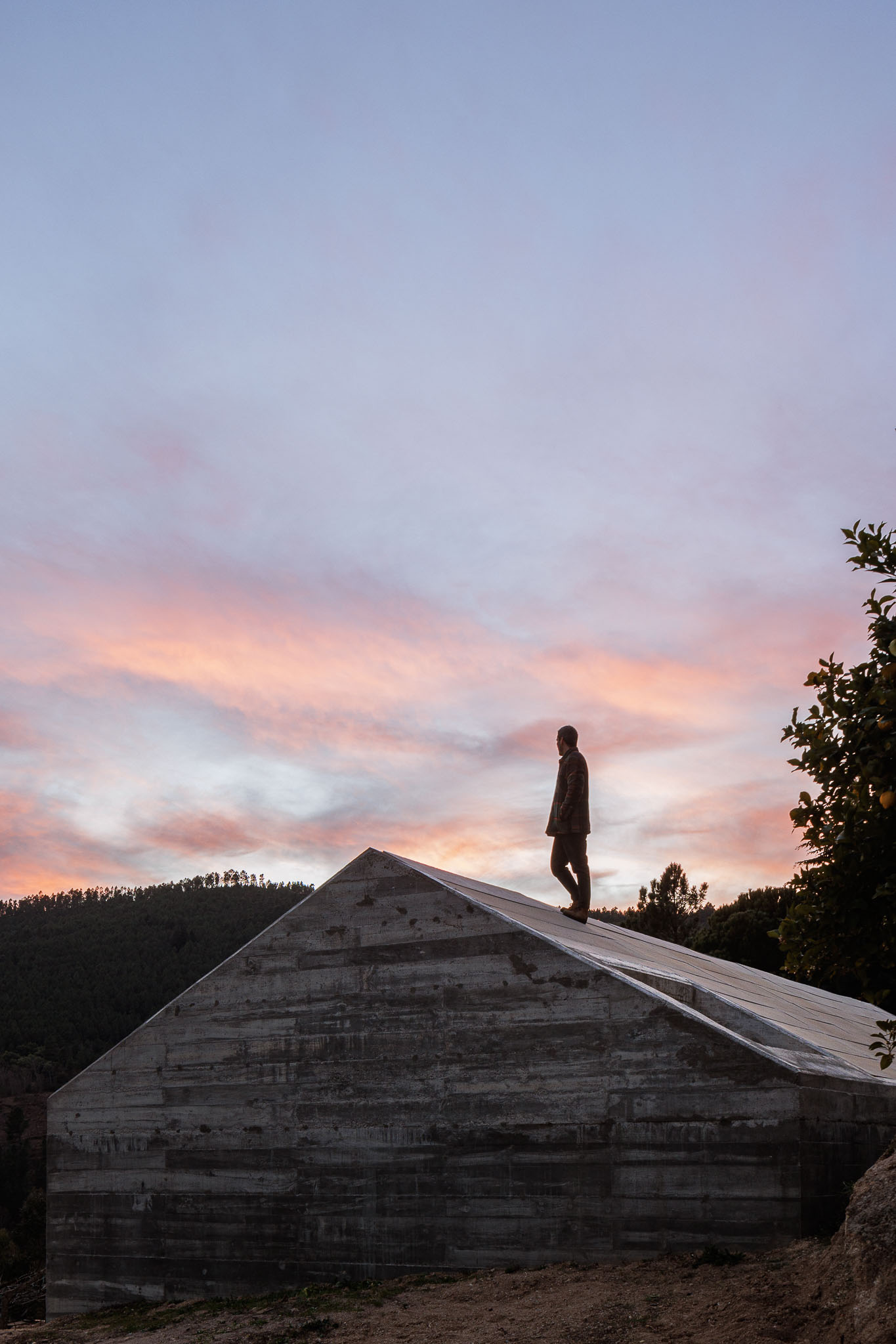 Casa NaMora: Silhueta de homem no telhado da casa em betão ao pôr do sol, com céu em tons de rosa e laranja. — Casa NaMora arquitetura David Bilo Arquitectos e Filipe Pina Arquitectura em Guarda