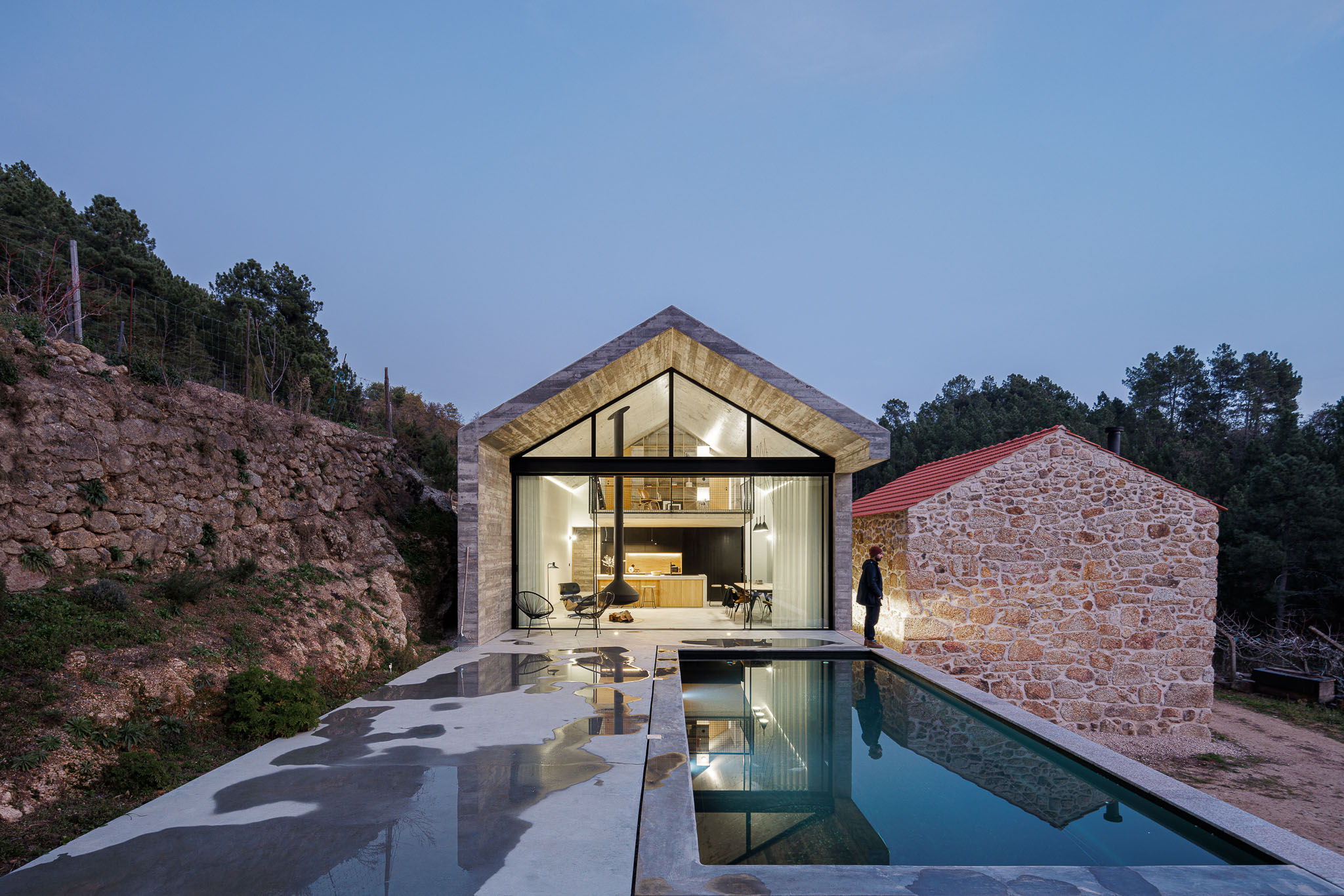 Casa NaMora: Fachada envidraçada da casa com vista para a sala de estar, piscina e anexo em pedra ao entardecer. — Casa NaMora arquitetura David Bilo Arquitectos e Filipe Pina Arquitectura em Guarda
