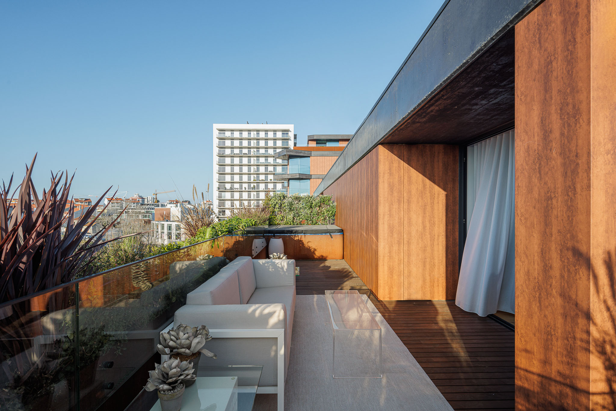Varanda com mobiliário branco, plantas, deck de madeira e vista para a cidade, com revestimento exterior em madeira. — Puro Homes arquitetura Mário Alves Arquitecto em Aveiro