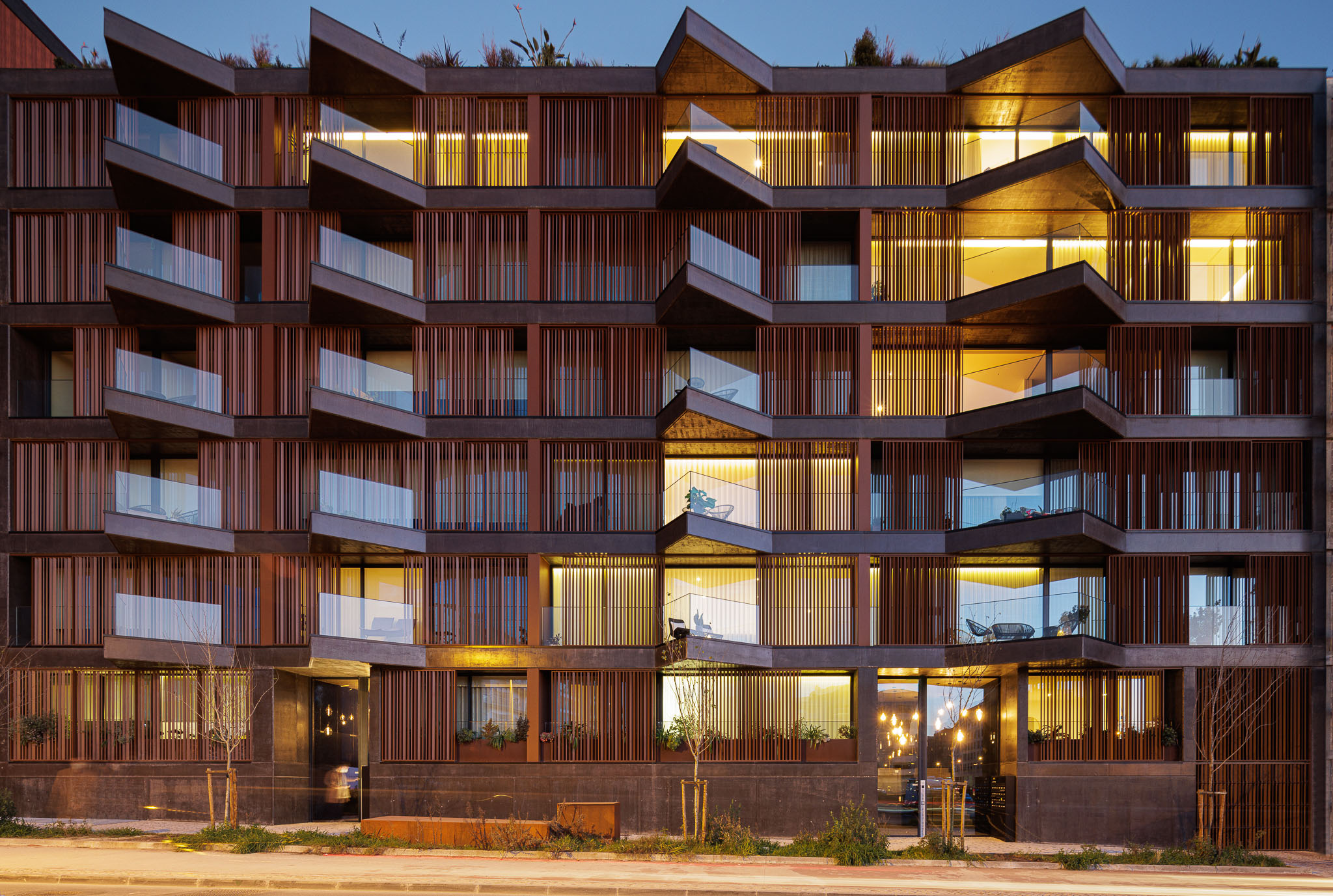 Edifício com fachada em madeira e betão, varandas angulares iluminadas e paisagismo urbano. — Puro Homes arquitetura Mário Alves Arquitecto em Aveiro