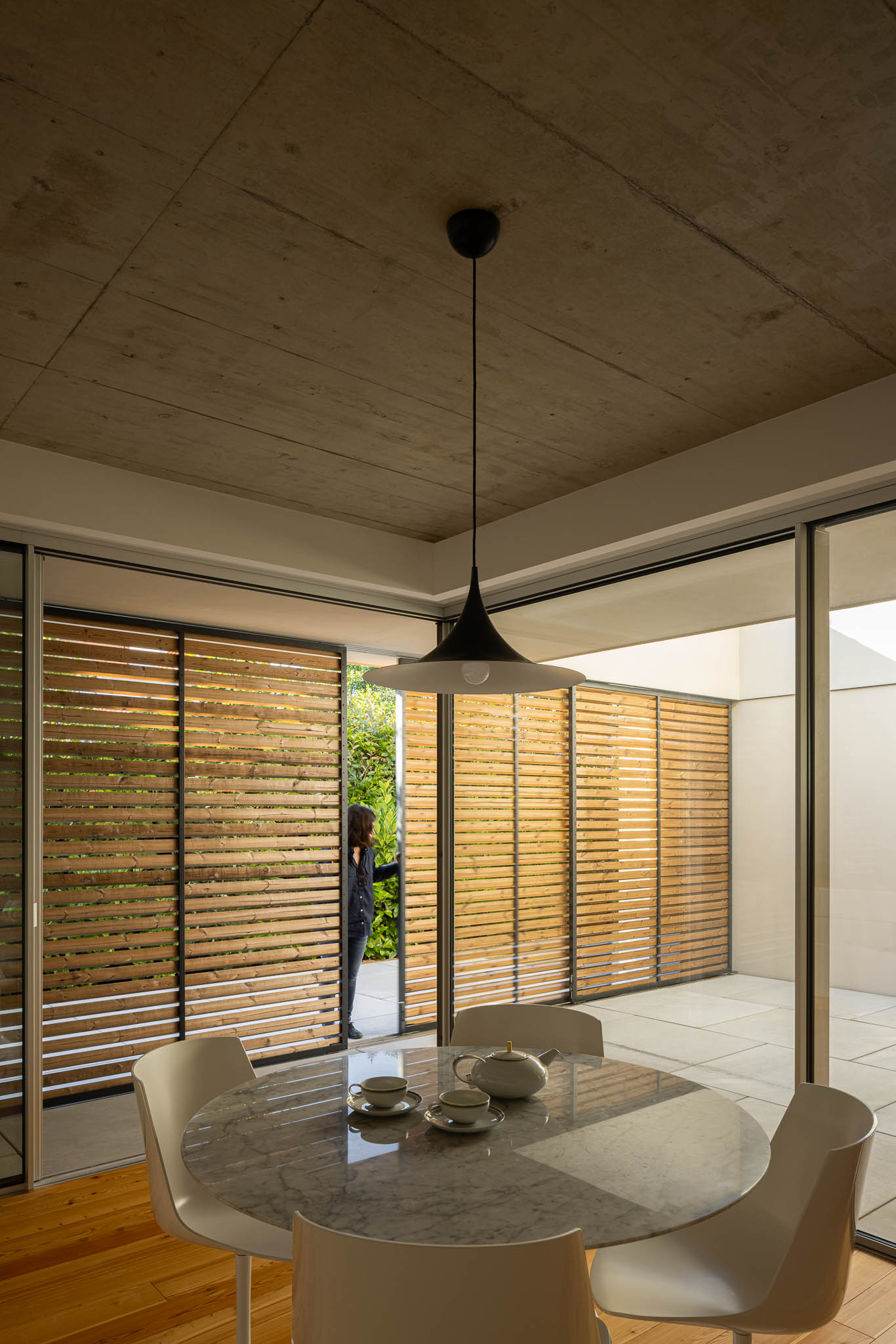 Sala de jantar: Mesa redonda em mármore, cadeiras brancas e candeeiro suspenso, com ripas de madeira. — Casa em Ílhavo arquitetura M2Senos Arquitectos em Ílhavo