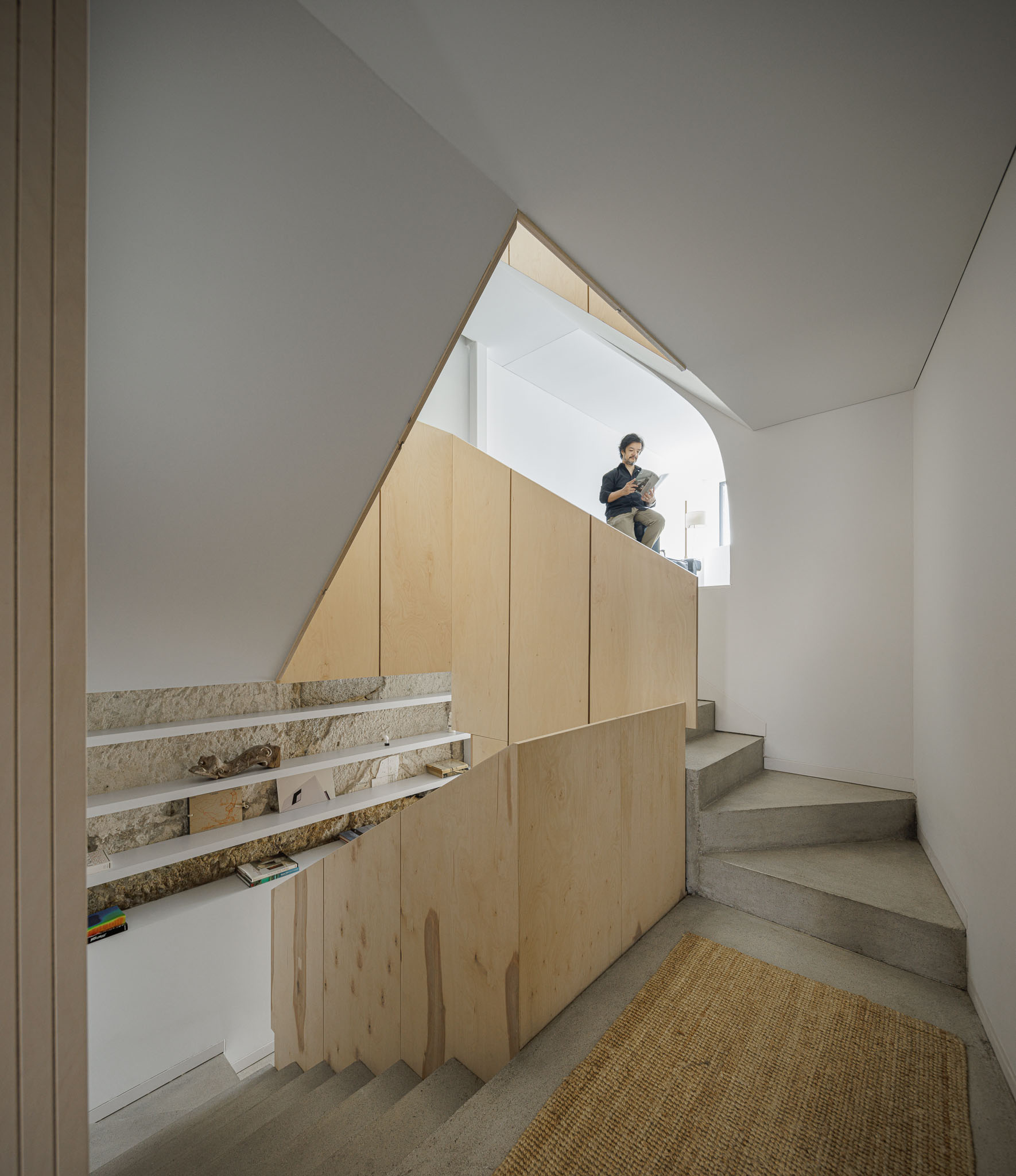 Escada: Corrimão em contraplacado claro, degraus em betão, nichos com prateleiras e alcatifa de juta. Pessoa a ler junto à janela. — Casa Vertical arquitetura Tsou Arquitectos em Porto