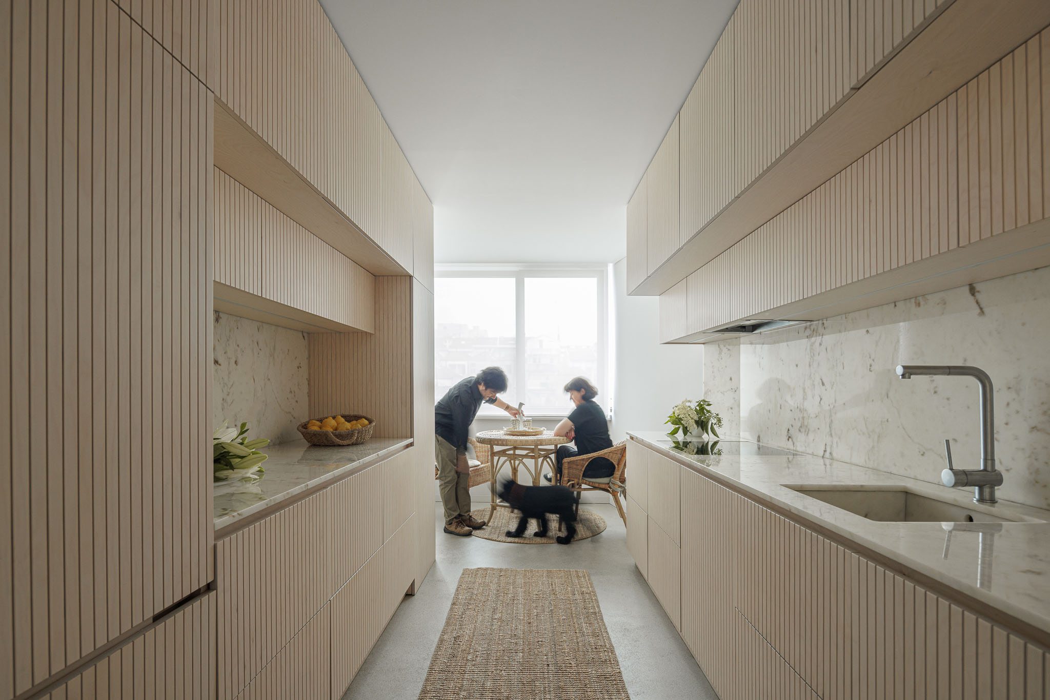Cozinha: Armários em madeira clara canelada, bancada em mármore, pavimento em betão afagado, mesa de refeições redonda em vime. — Casa Vertical arquitetura Tsou Arquitectos em Porto