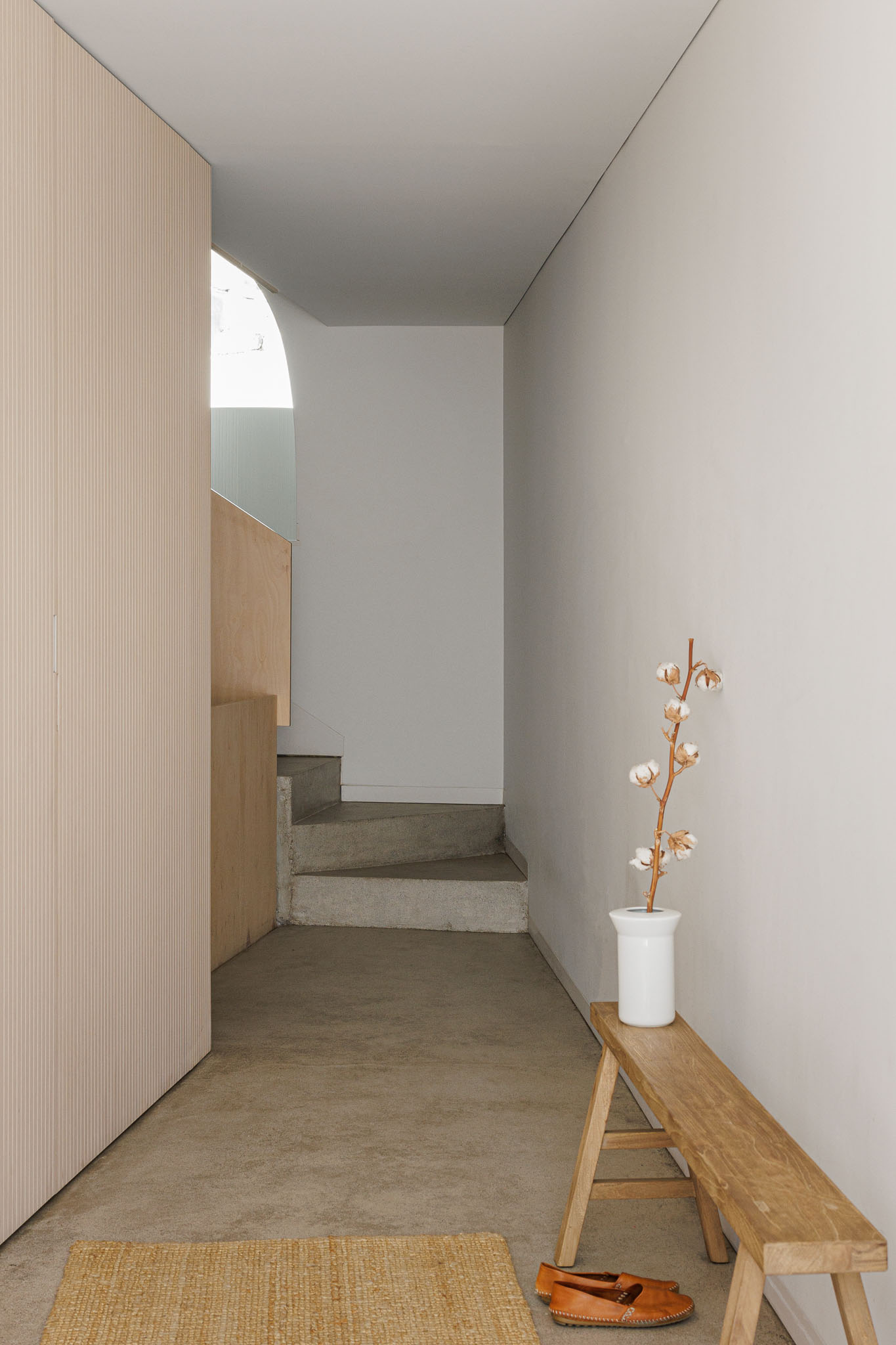 Hall de entrada: Pavimento em betão afagado, banco de madeira rústico, vaso com flor de algodão, porta em madeira clara canelada. — Casa Vertical arquitetura Tsou Arquitectos em Porto
