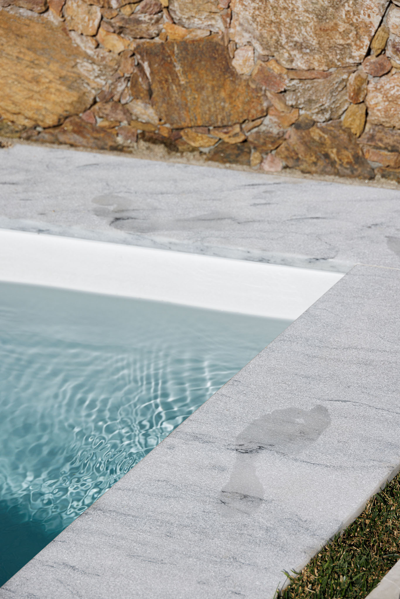Piscina: Detalhe da borda em mármore cinzento e água azul, com parede em pedra ao fundo. — Casa Forte arquitetura pema studio em Santo Tirso