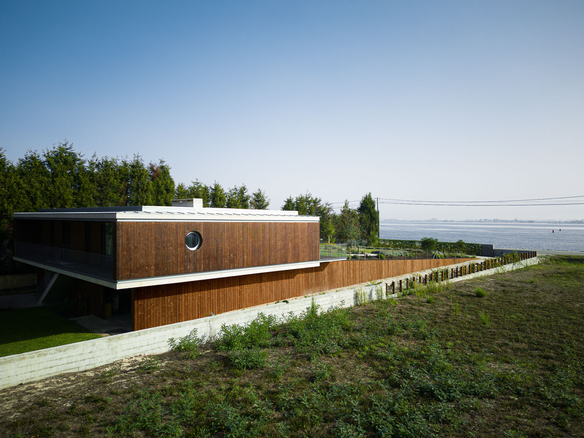 Vista lateral da casa com revestimento em madeira, janela circular e acesso à ria. — Casa na Torreira arquitetura Nu.ma em Torreira