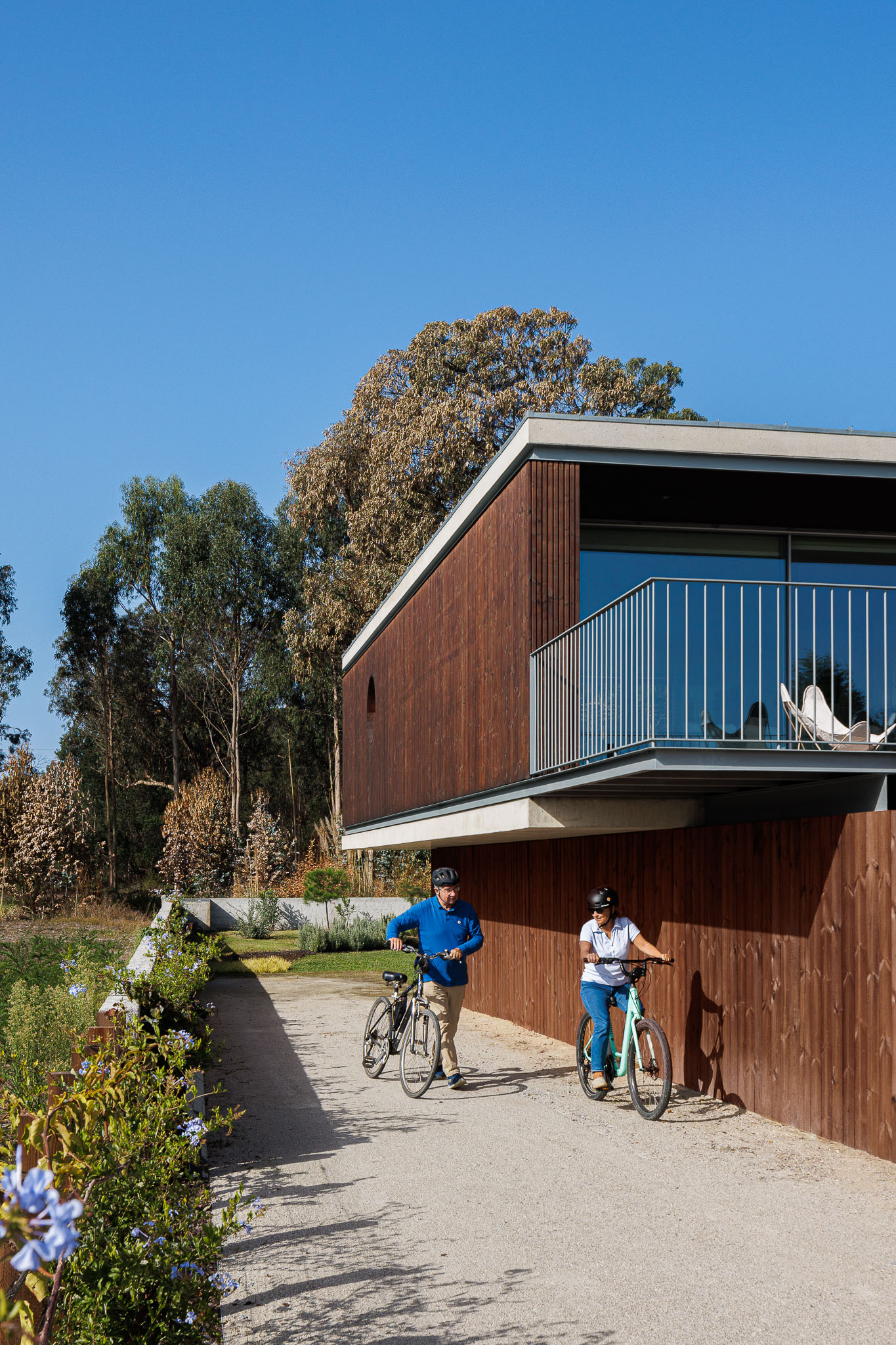 Detalhe da fachada em madeira da casa, varanda com gradeamento metálico e pessoas com bicicletas. — Casa na Torreira arquitetura Nu.ma em Torreira