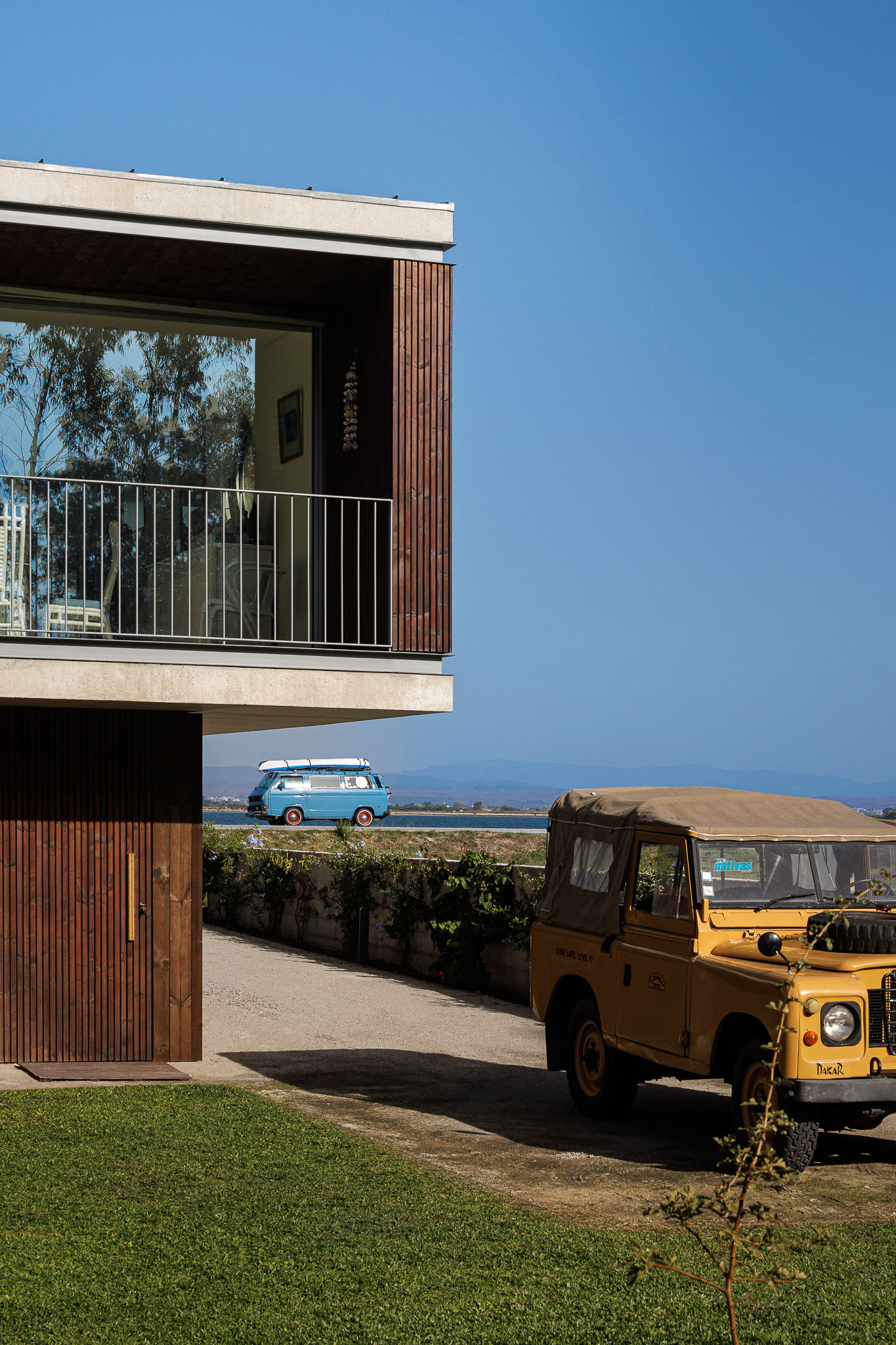 Detalhe da fachada em madeira, varanda com gradeamento metálico e carros estacionados. — Casa na Torreira arquitetura Nu.ma em Torreira