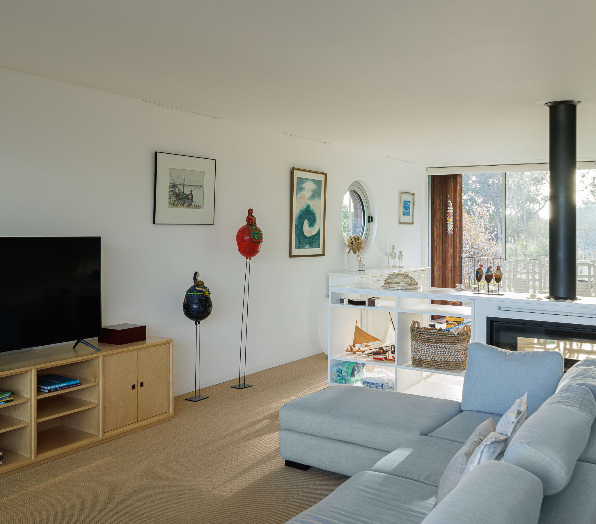 Sala de estar com sofá azul claro, lareira, móveis de madeira clara e vista para o exterior. — Casa na Torreira arquitetura Nu.ma em Torreira