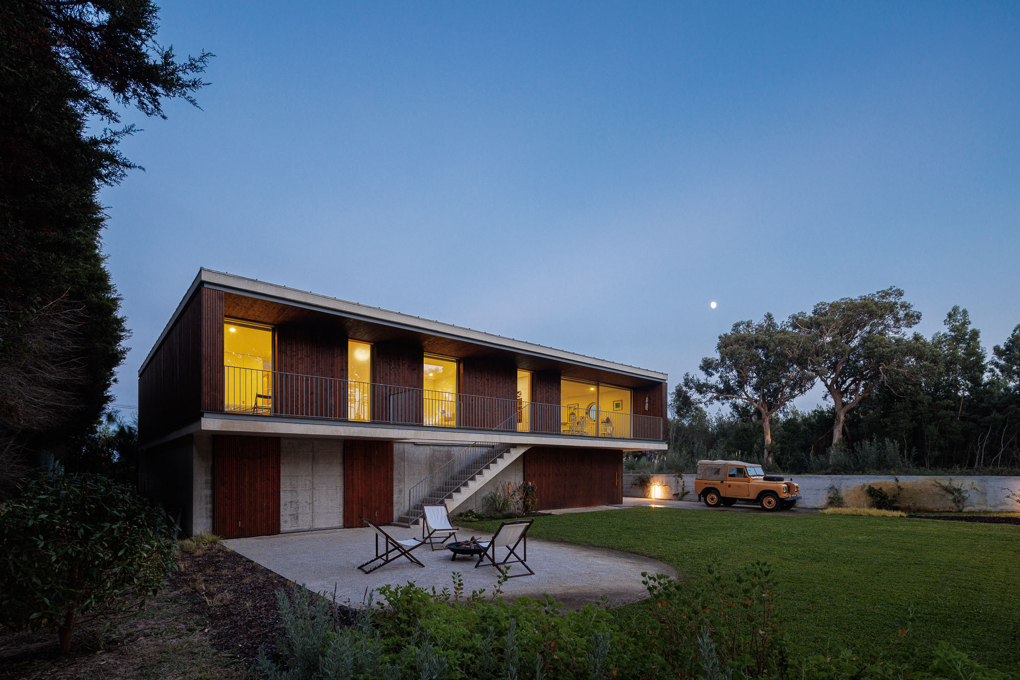 Exterior da casa à noite, com iluminação interior e exterior, jardim relvado e árvores. — Casa na Torreira arquitetura Nu.ma em Torreira