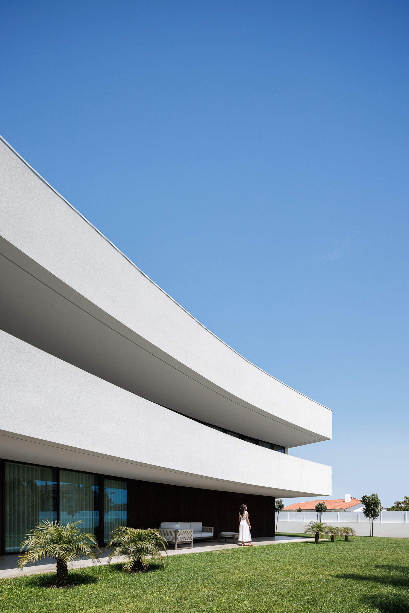 Jardim: Fachada branca curva da casa, com relvado, palmeiras e mobiliário de exterior minimalista. — Casa 109 arquitetura Frari em Vagos