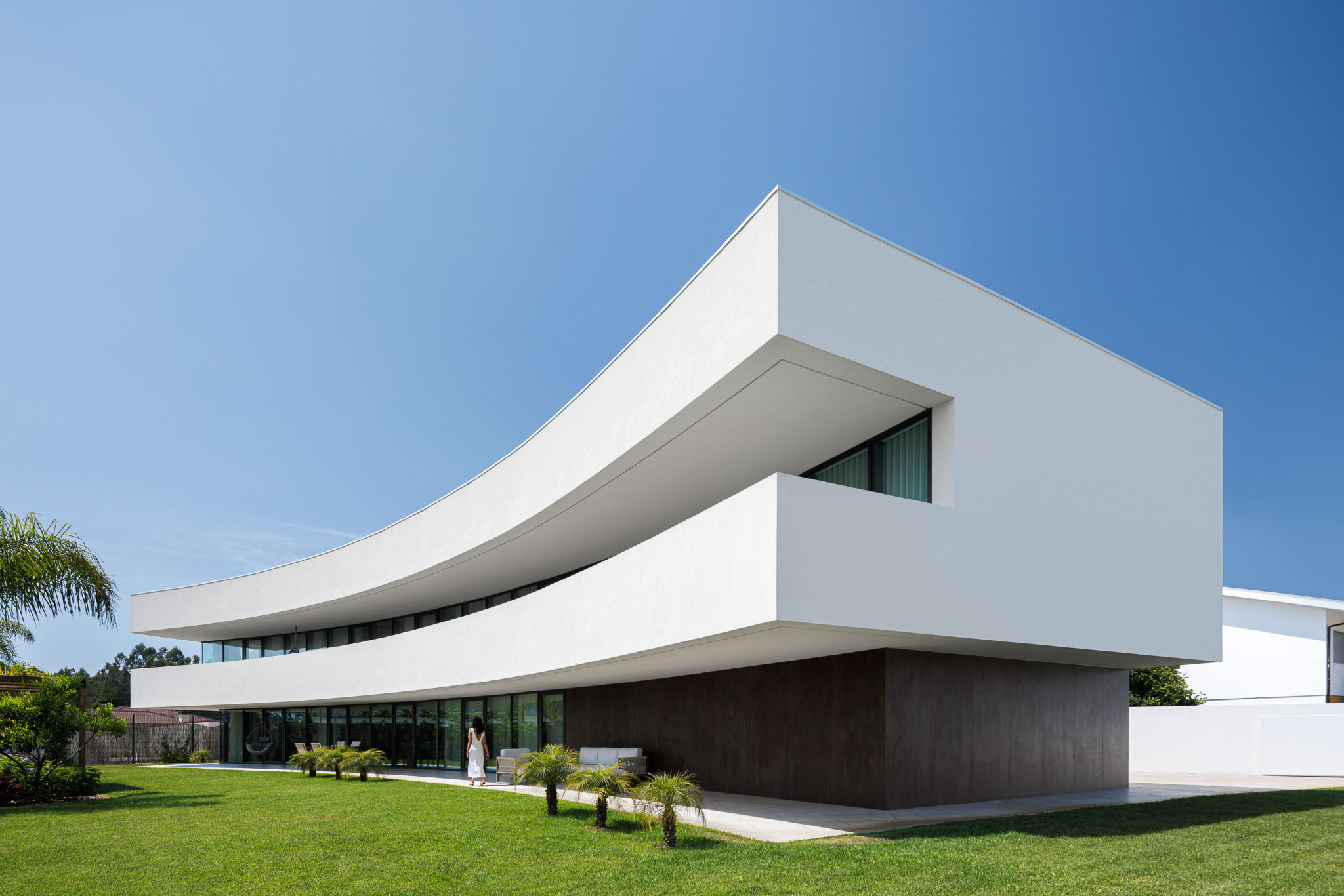Exterior: Vista angular da casa de dois pisos com design curvo e acabamentos em branco e castanho. — Casa 109 arquitetura Frari em Vagos