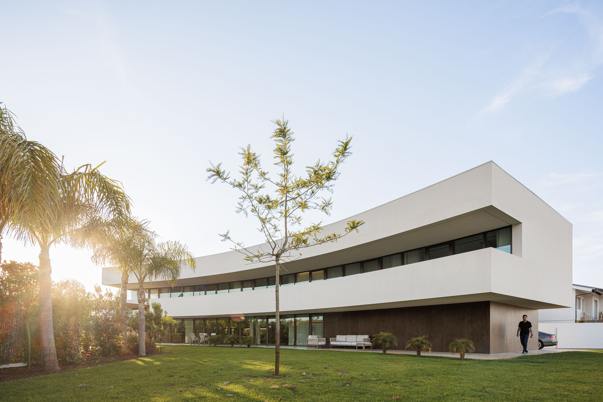 Exterior: Vista da casa com relvado, palmeiras e fachada branca curva com detalhes em castanho. — Casa 109 arquitetura Frari em Vagos