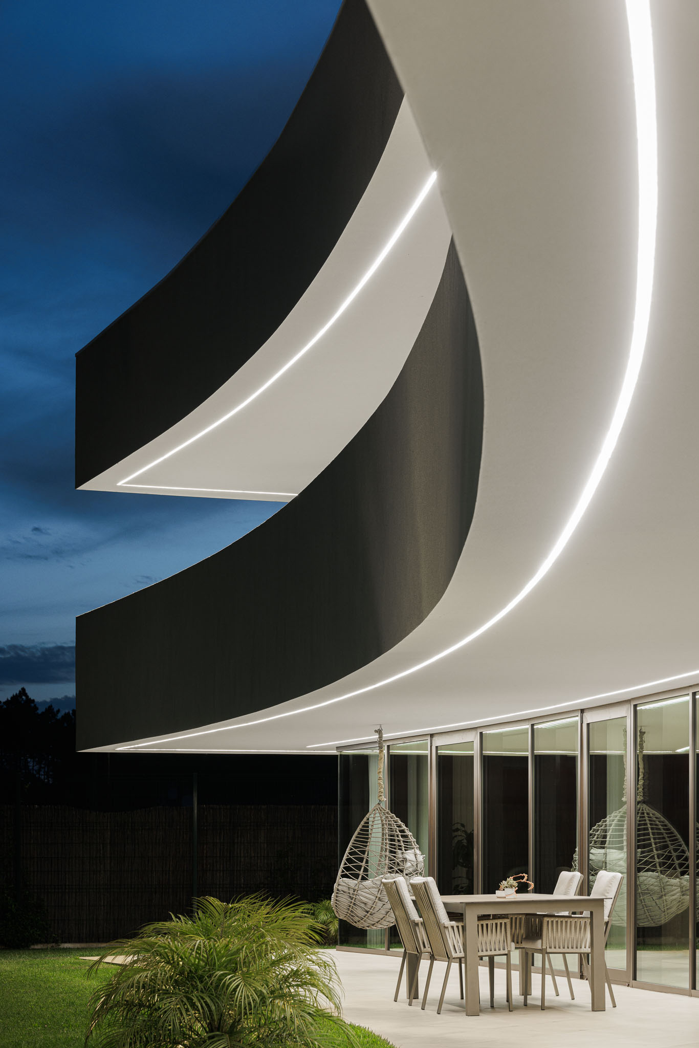 Alpendre Noturno: Mesa de jantar e cadeiras no alpendre iluminado, com cadeiras suspensas e vista para o jardim. — Casa 109 arquitetura Frari em Vagos