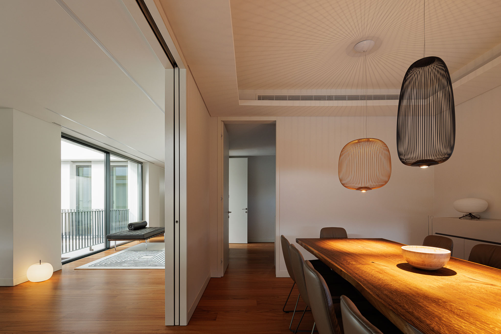 Sala de jantar: Vista da sala de jantar para a sala de estar, com mesa de madeira e candeeiros suspensos. — CONCRETE 11 arquitetura Cage Atelier em Lisboa