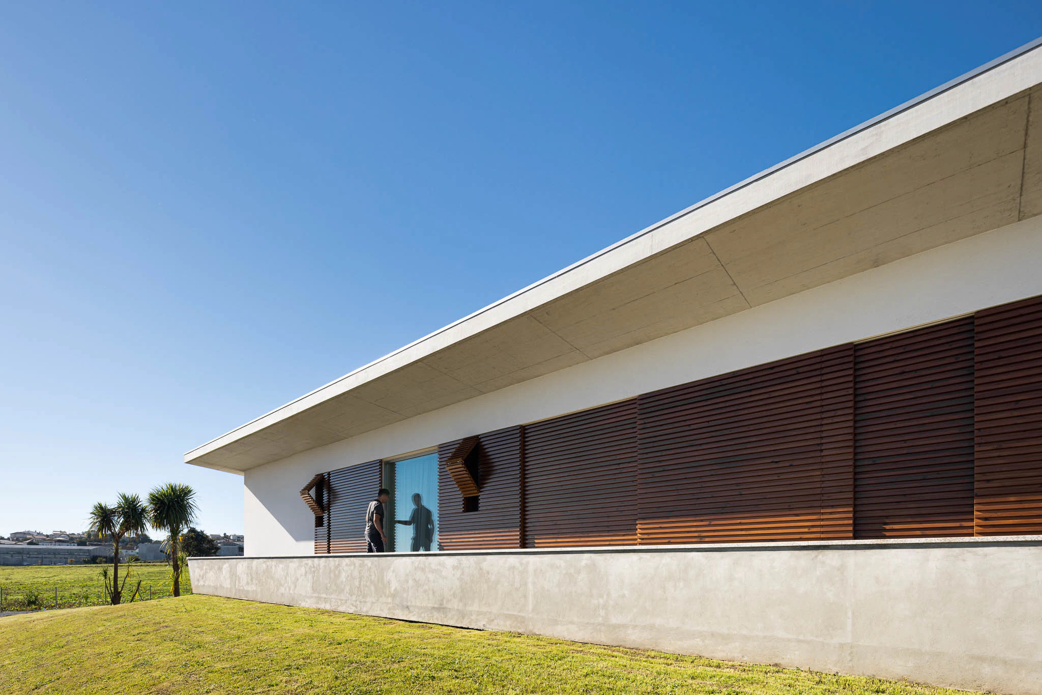 Fachada minimalista com ripas de madeira e detalhes geométricos, sob céu azul e relvado verde. — Casa em Brufe arquitetura Oval em Vila Nova de Famalicão