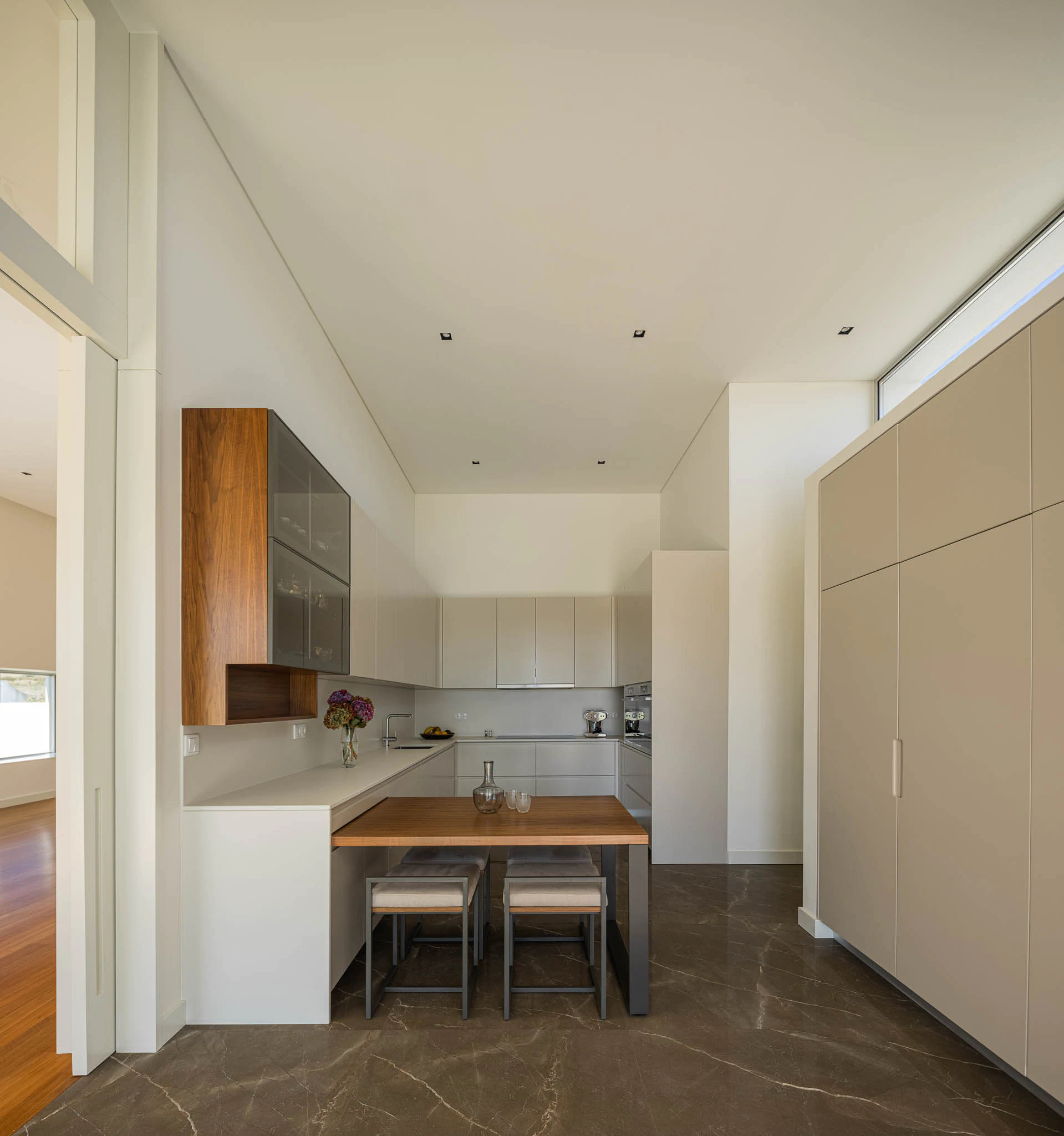 Cozinha moderna com armários brancos e detalhes em madeira, pavimento marmoreado e luz natural. — Casa em Brufe arquitetura Oval em Vila Nova de Famalicão
