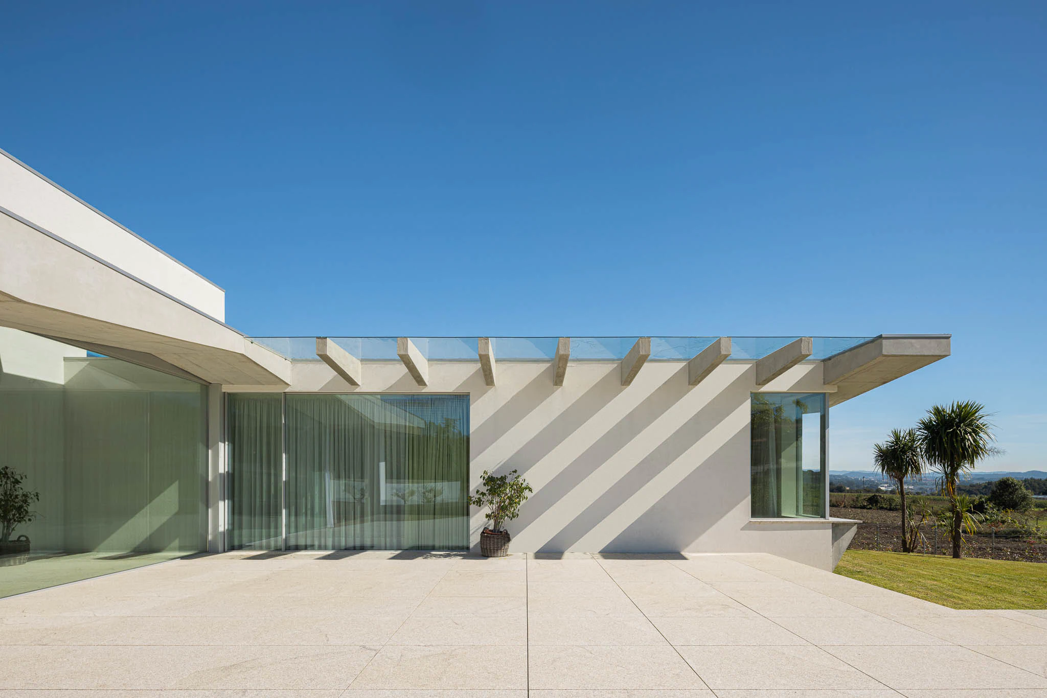 Alpendre com cobertura de vidro e vigas de betão, paredes brancas e pavimento em pedra clara. — Casa em Brufe arquitetura Oval em Vila Nova de Famalicão