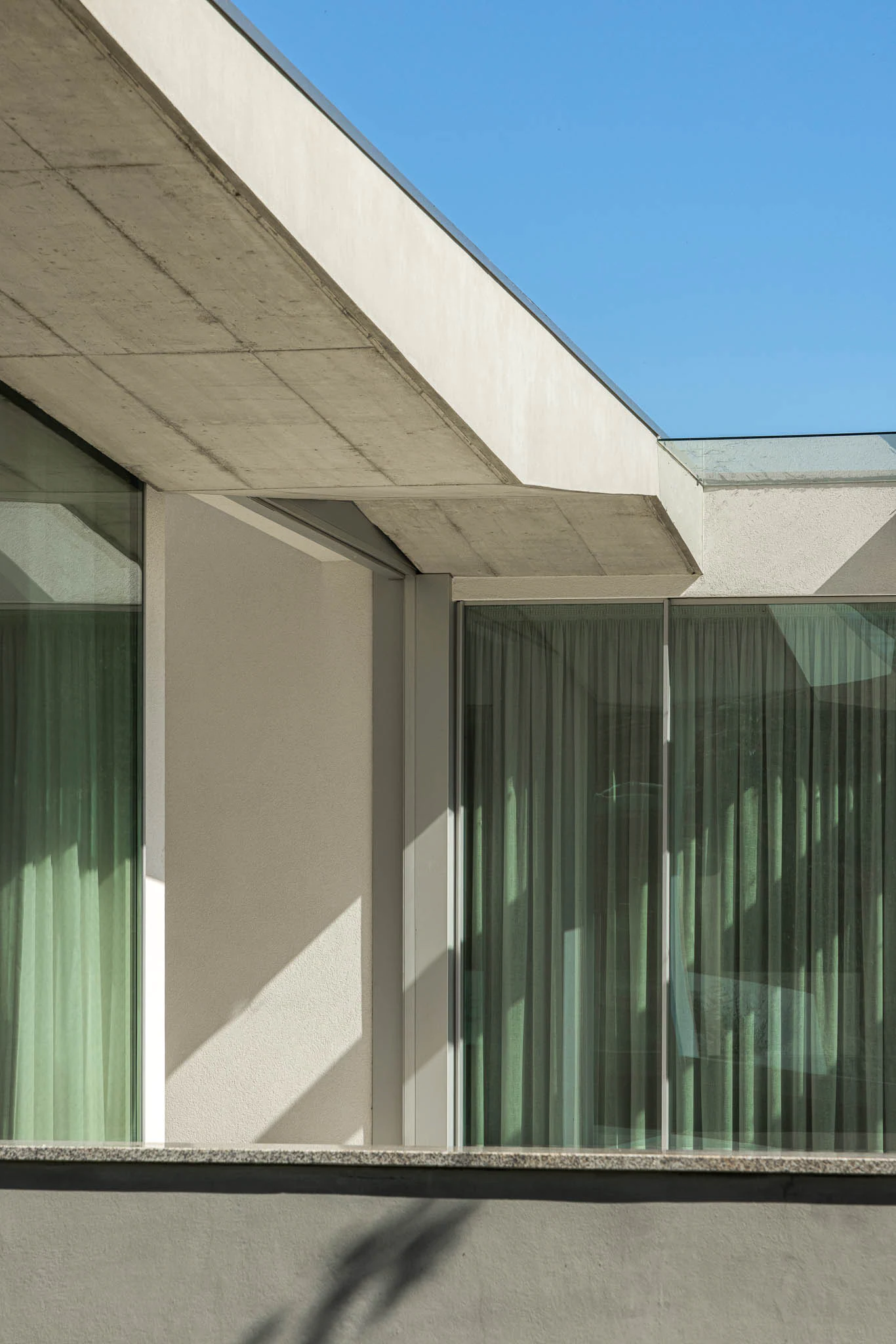Pormenor da fachada com cobertura de betão, janelas amplas e cortinas translúcidas. — Casa em Brufe arquitetura Oval em Vila Nova de Famalicão
