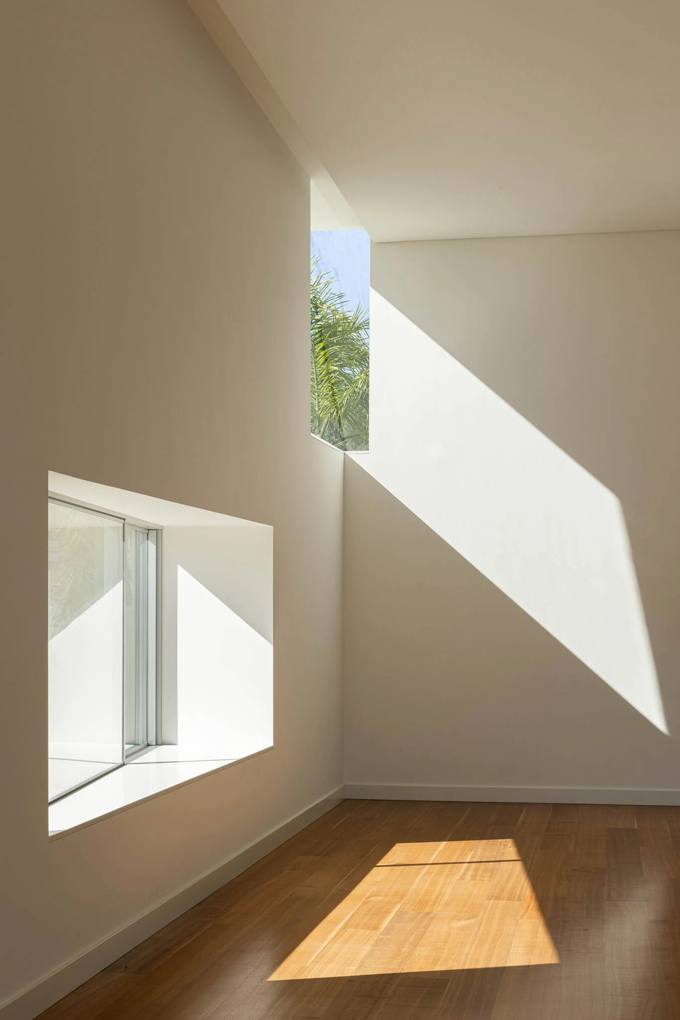 Interior com janelas geométricas, paredes brancas e chão em madeira, com luz natural intensa. — Casa em Brufe arquitetura Oval em Vila Nova de Famalicão