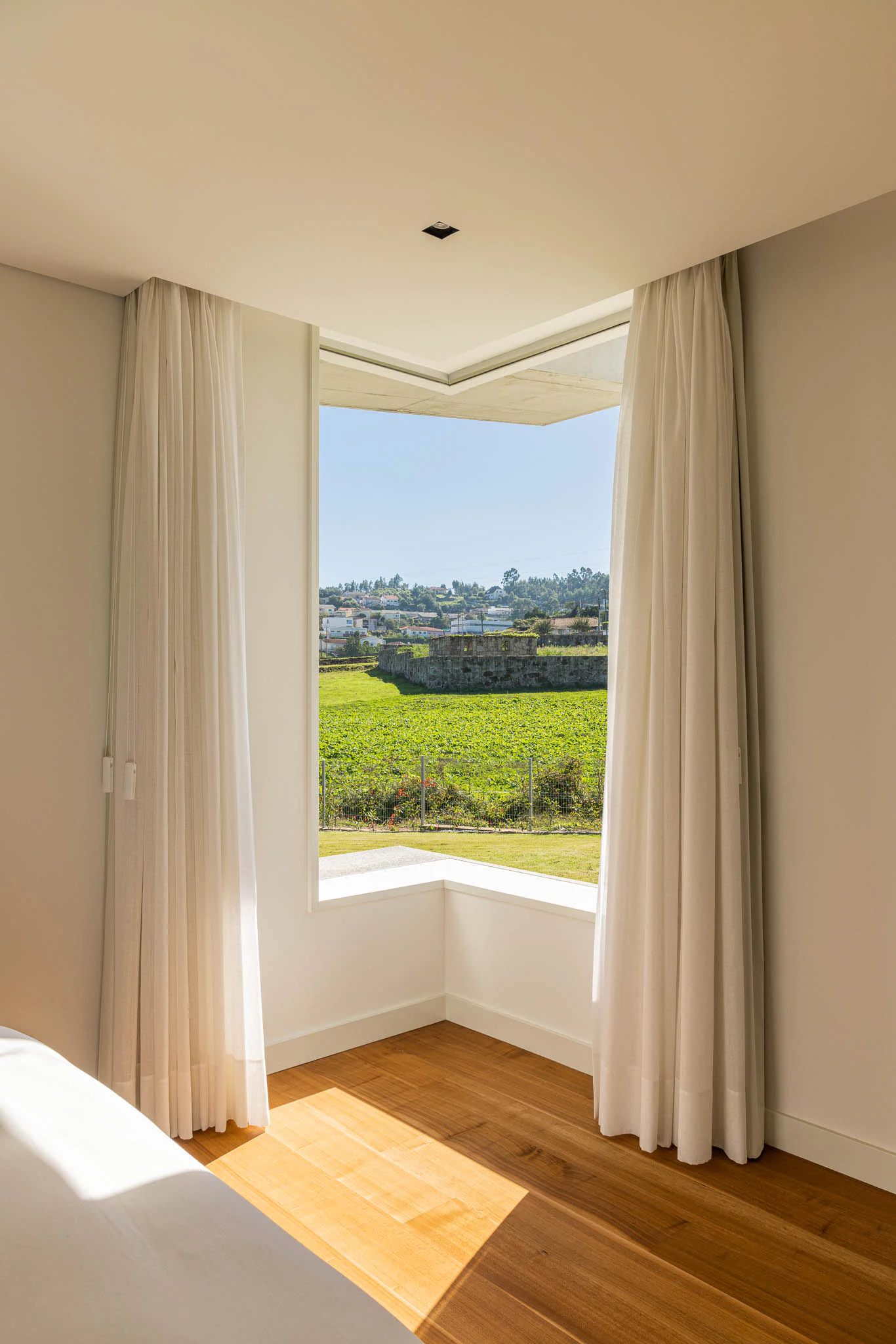Quarto com vista para o campo, cortinas brancas e chão em madeira, com luz suave. — Casa em Brufe arquitetura Oval em Vila Nova de Famalicão