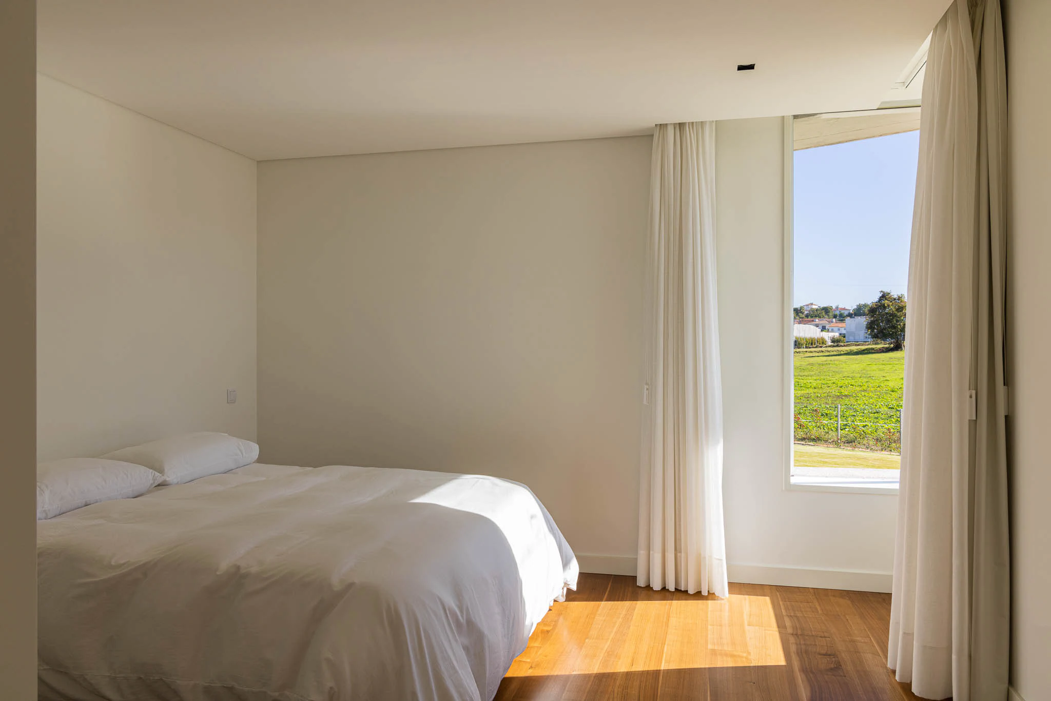 Quarto com cama de casal, cortinas brancas e vista para o exterior, com luz natural. — Casa em Brufe arquitetura Oval em Vila Nova de Famalicão