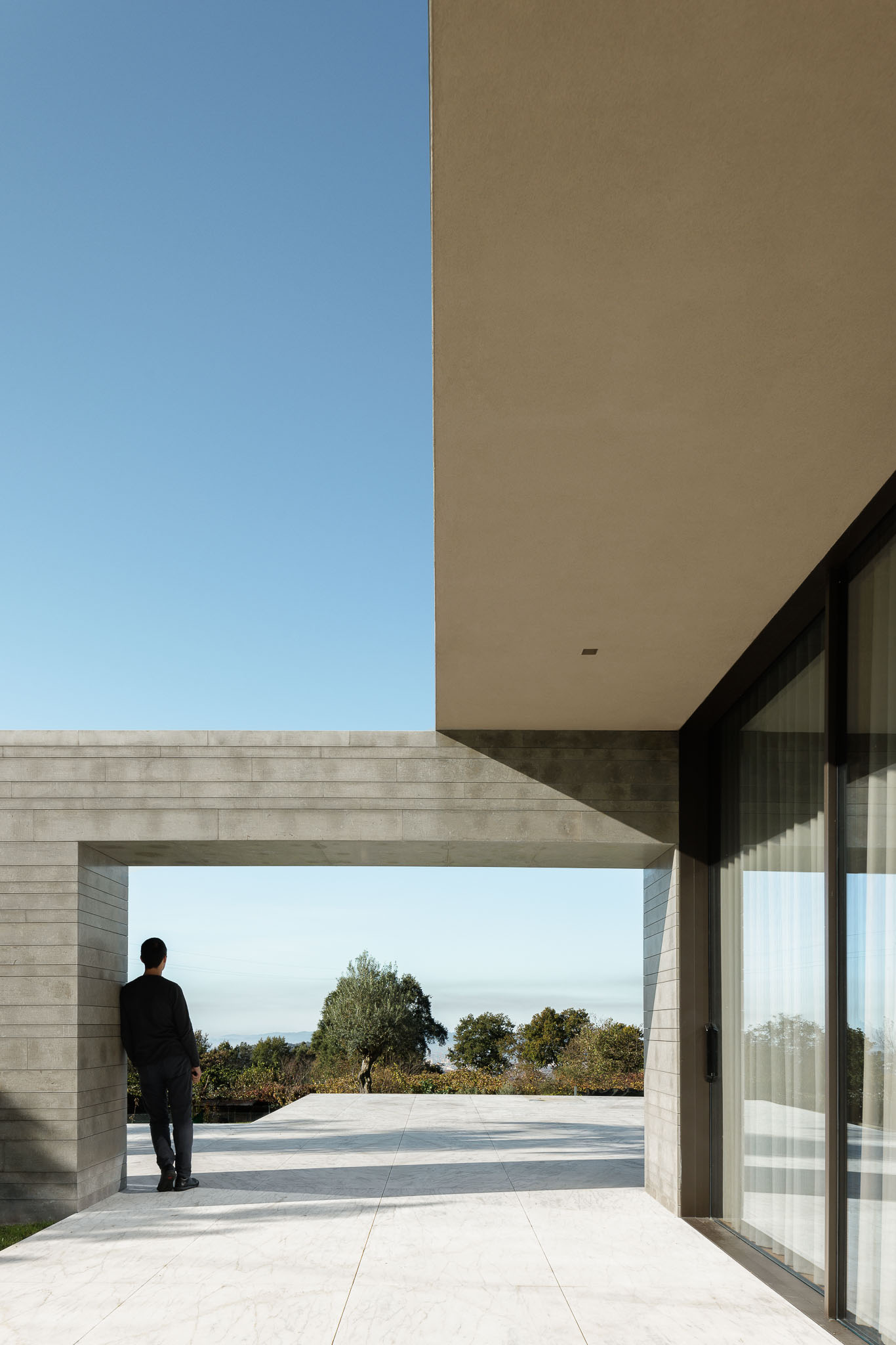 Alpendre: Homem encostado à parede de betão do alpendre com pavimento em mármore e vista para a paisagem. — Casa D arquitetura L2C Arquitetura em Braga