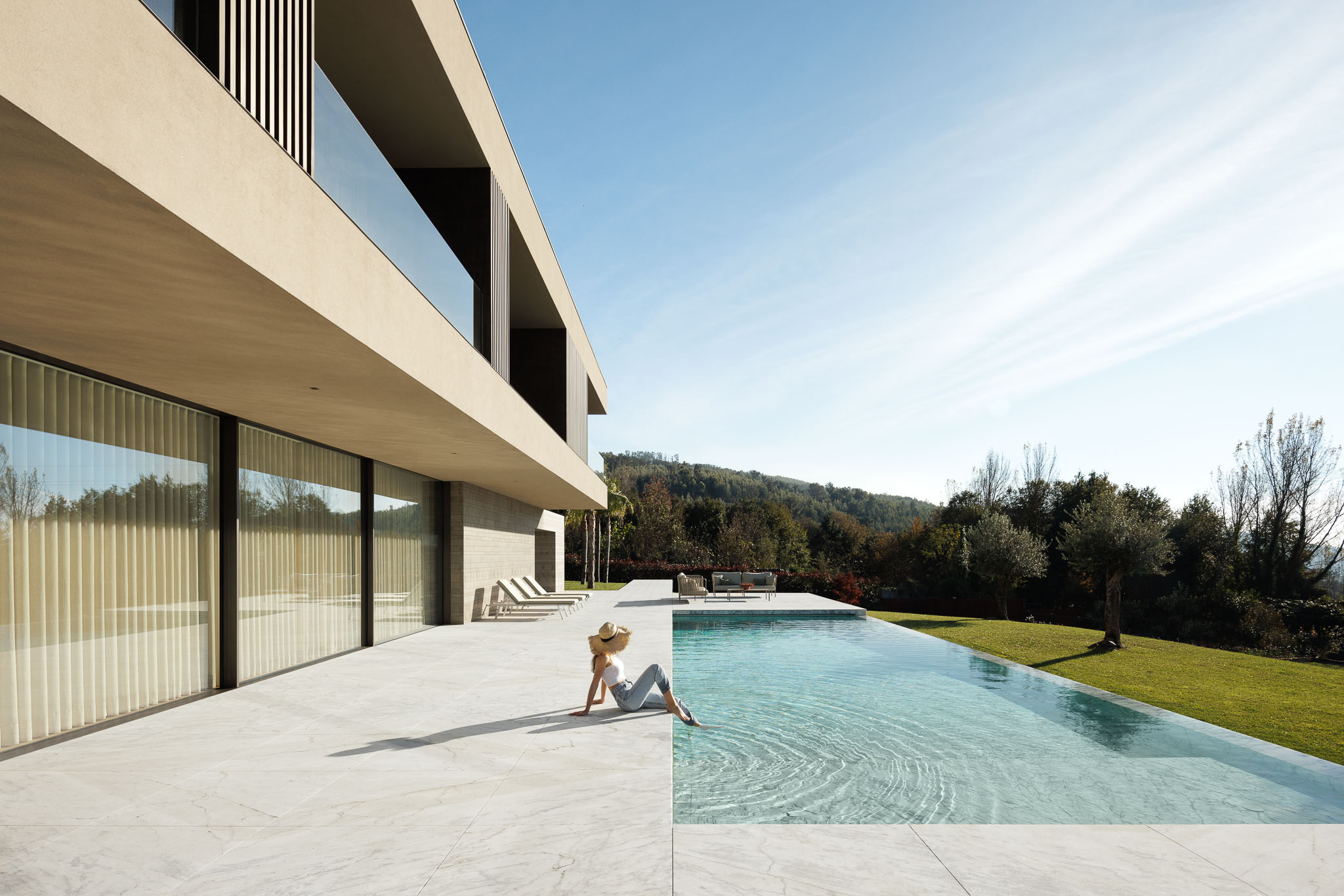Piscina: Mulher sentada na borda da piscina infinita com pavimento em mármore e vista para a paisagem. — Casa D arquitetura L2C Arquitetura em Braga