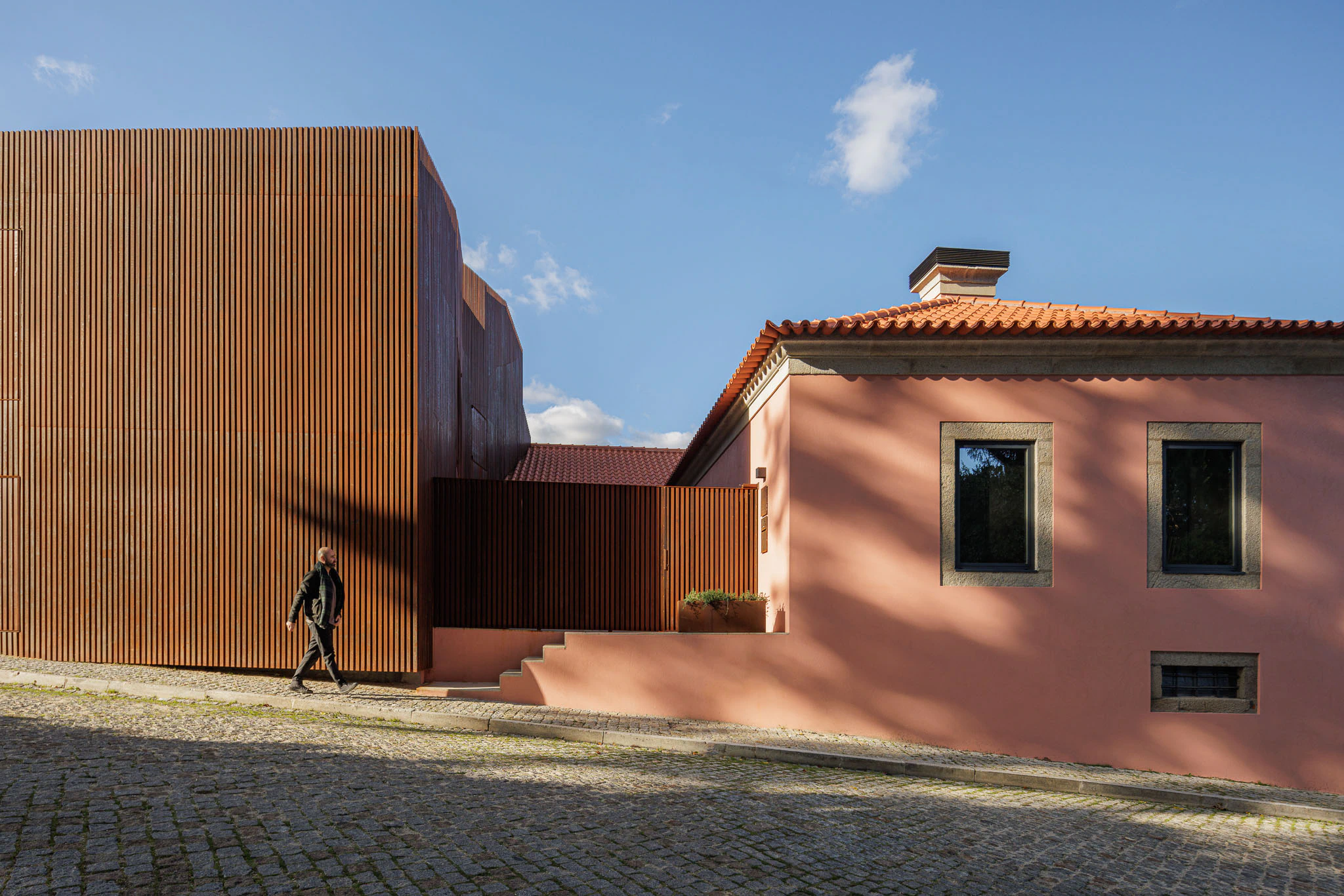 Casa da Trincheira: Fachada da casa cor-de-rosa e anexo de a&ccedil;o corten, com um homem a caminhar na rua de cal&ccedil;ada. &mdash; Casa da Trincheira arquitetura David Bilo Arquitectos e Filipe Pina Arquitectura em Pinhel