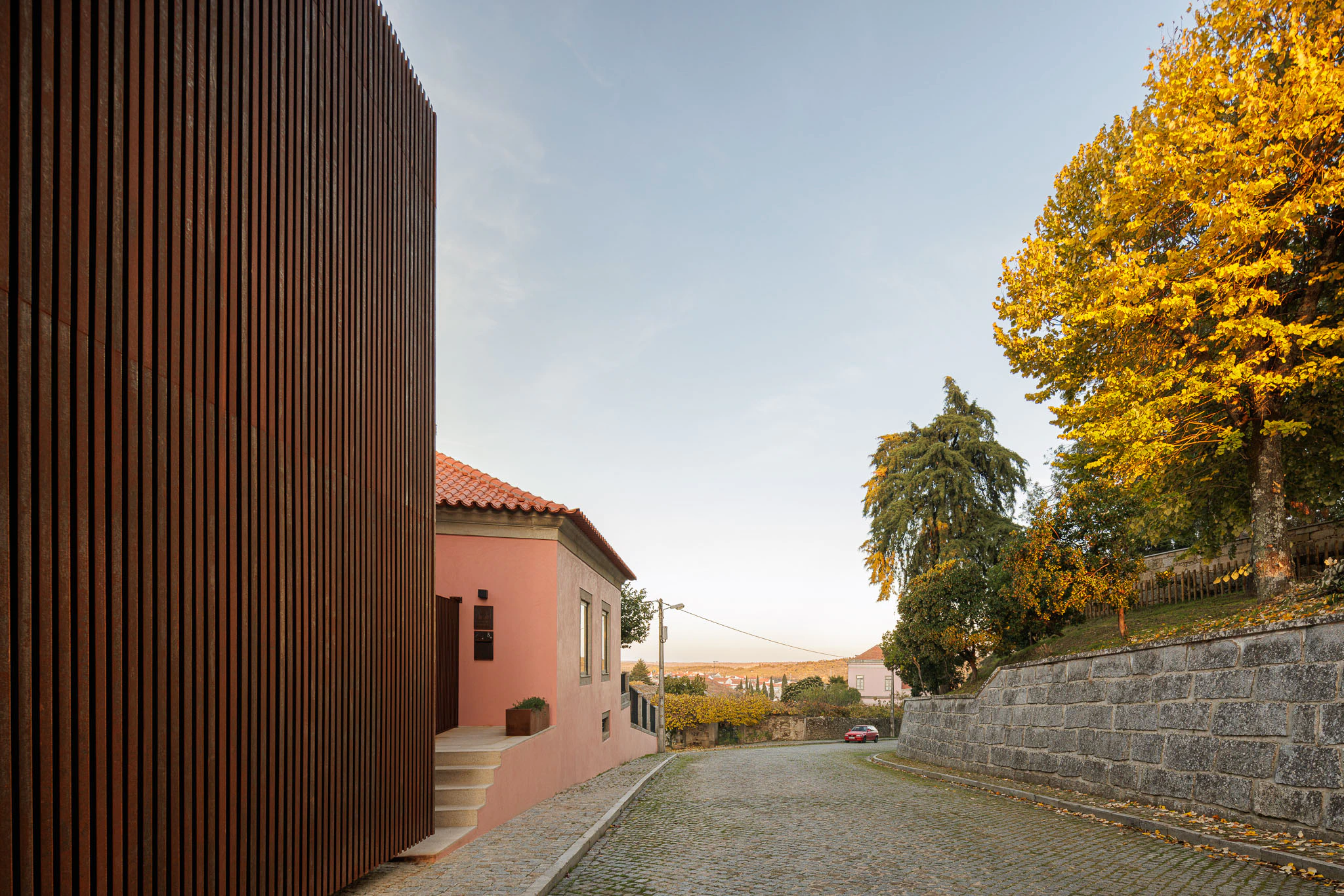 Casa da Trincheira: Perspectiva da rua com a casa cor-de-rosa e o anexo em aço corten, integrados na paisagem urbana. — Casa da Trincheira arquitetura David Bilo Arquitectos e Filipe Pina Arquitectura em Pinhel