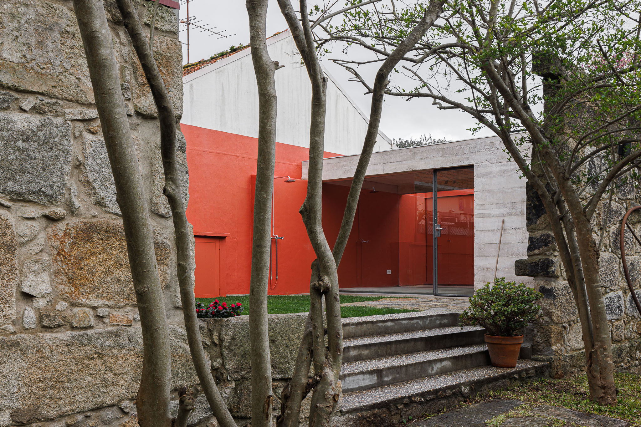 Entrada: Fachada moderna em betão e vermelho, com porta envidraçada, vista através de árvores. — Pavilhão de jardim arquitetura José Pedro Lima Arquiteto em Porto