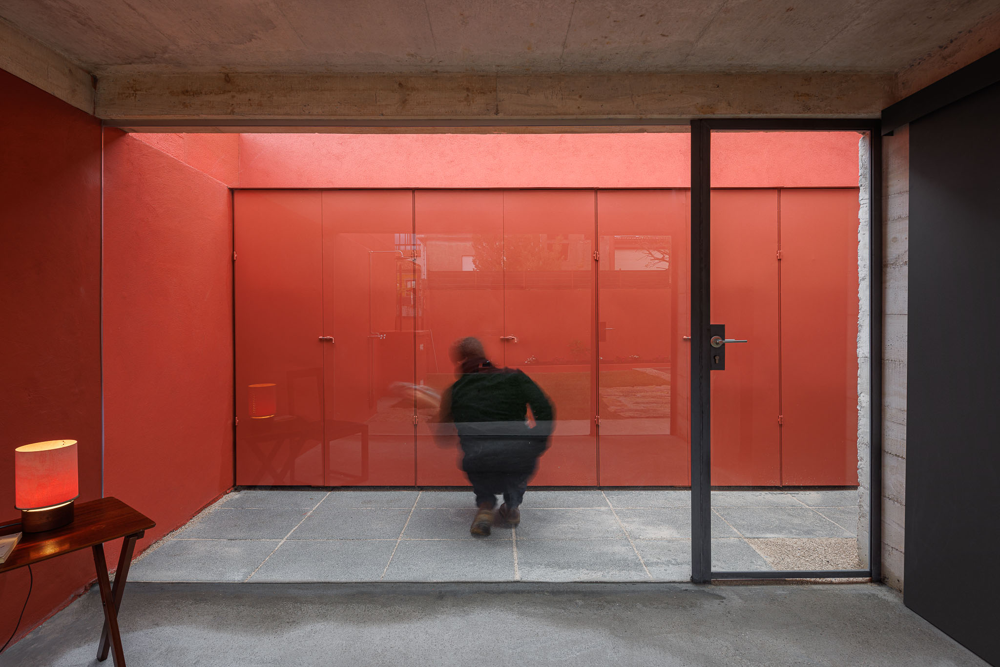 Interior: Espaço com pavimento cinzento, paredes vermelhas, candeeiro e pessoa a passar pela porta envidraçada. — Pavilhão de jardim arquitetura José Pedro Lima Arquiteto em Porto