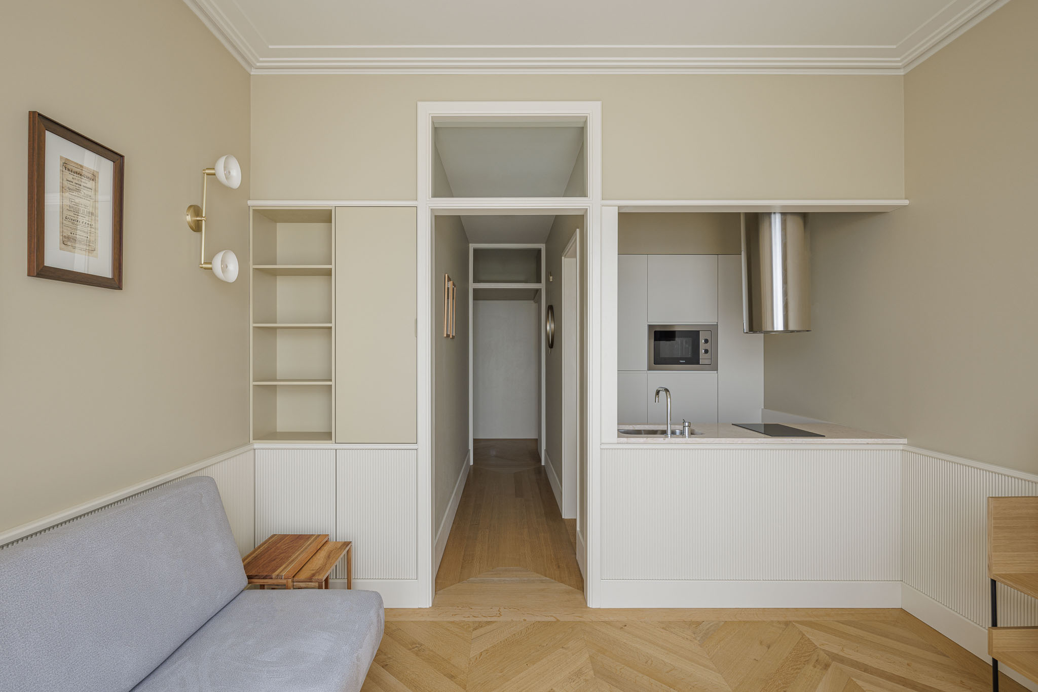 Sala de estar: Sofá cinzento, estantes embutidas e acesso à cozinha, com pavimento em madeira clara e paredes bege. — Villa Theatro arquitetura arquitectos aliados em Braga