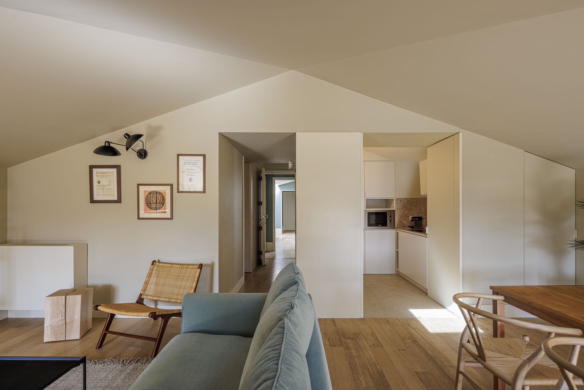 Sala de estar: Sofá azul, cadeirão de vime e acesso à cozinha, com pavimento em madeira clara e teto inclinado. — Villa Theatro arquitetura arquitectos aliados em Braga