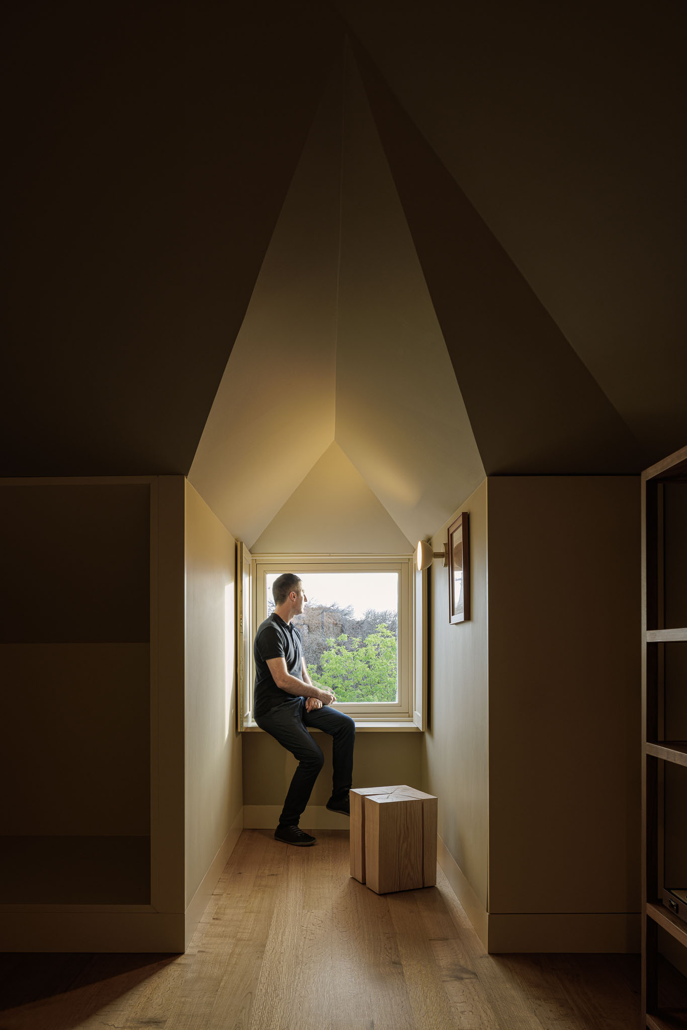Quarto: Pessoa sentada junto à janela com teto inclinado, com pavimento em madeira clara e paredes bege. — Villa Theatro arquitetura arquitectos aliados em Braga