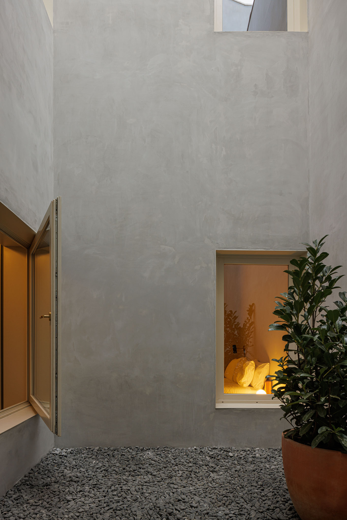 Pátio interior: Pátio com seixos no pavimento, paredes em betão aparente e janelas, com planta em vaso. — Villa Theatro arquitetura arquitectos aliados em Braga