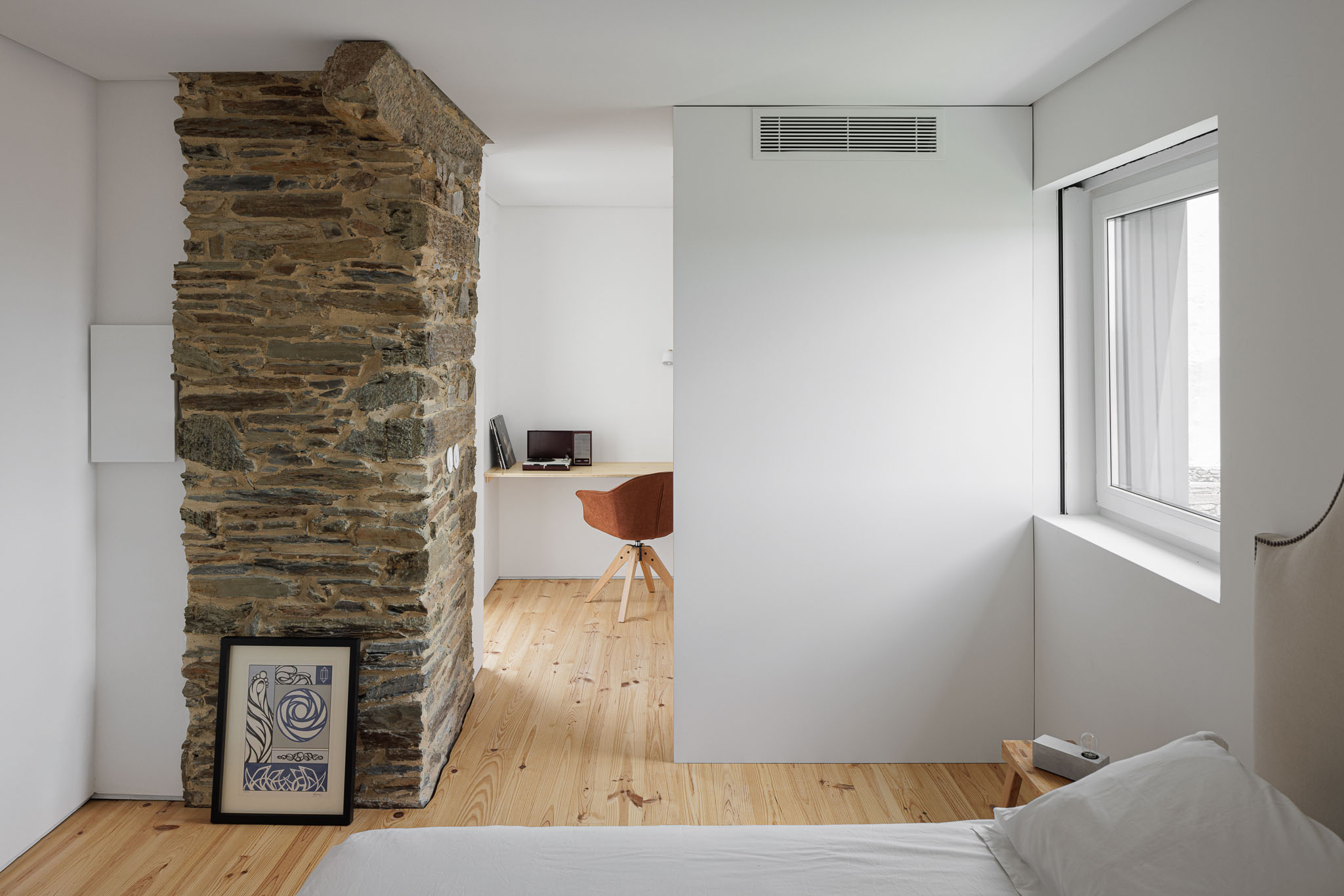 Quarto: Parede em pedra rústica divide quarto de escritório. Pavimento em madeira clara e paredes brancas. — Casa Caldeira arquitetura Filipe Pina Arquitectura em Vila Nova de Foz Côa