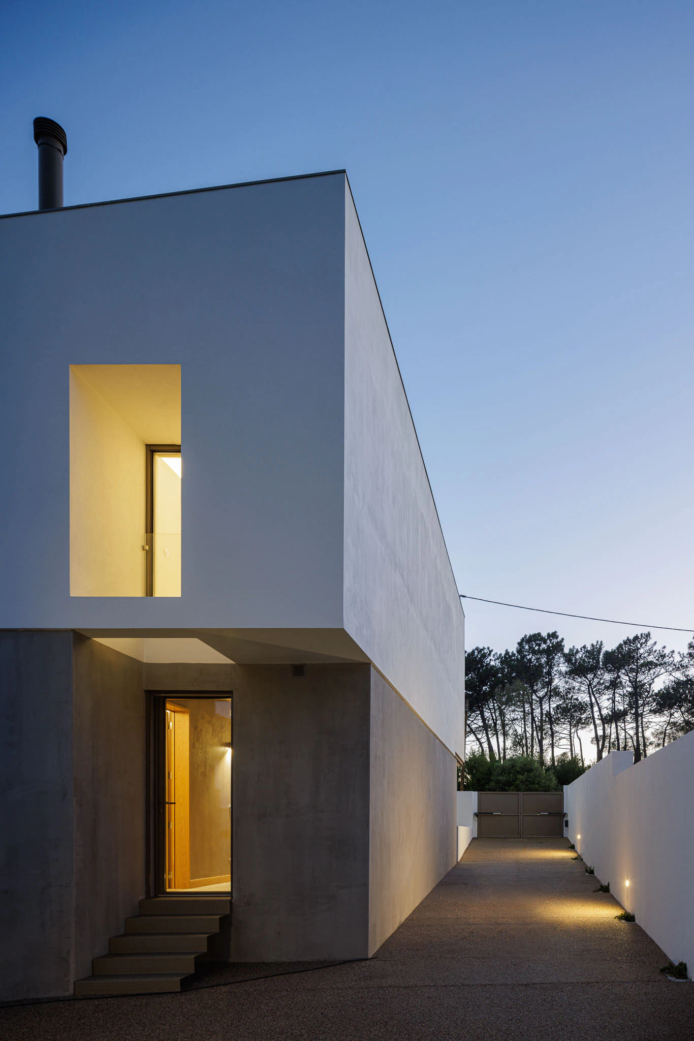 Fachada da casa iluminada com volume branco sobre base cinzenta, janela quadrada e porta de entrada. — Casas do Pinhal arquitetura Pk Arquitetos em Estoril