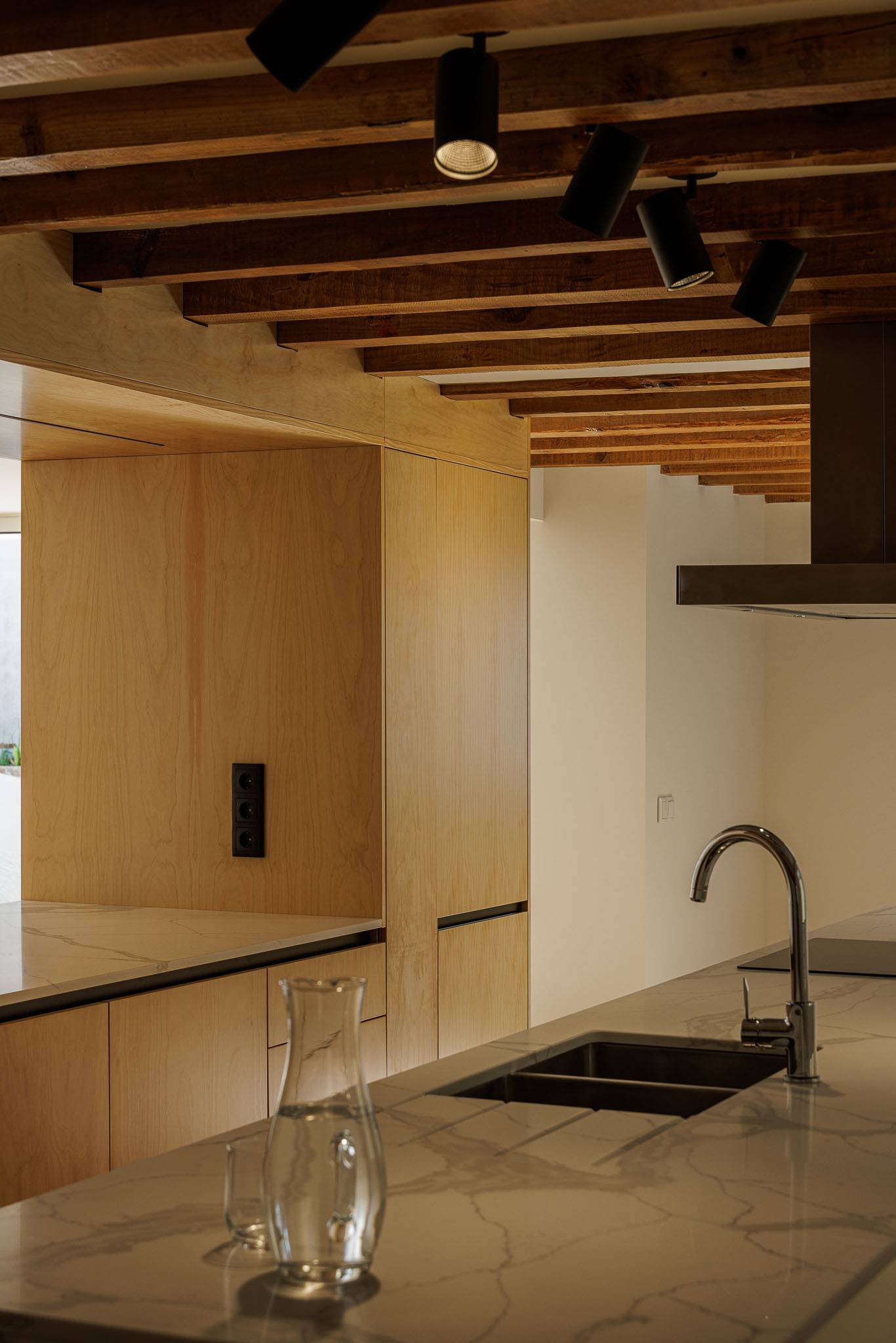 Cozinha: Detalhe da ilha da cozinha com armários em madeira clara, teto com vigas de madeira aparente e candeeiros pretos. — Casa dos anos 40 arquitetura Frari em Ílhavo