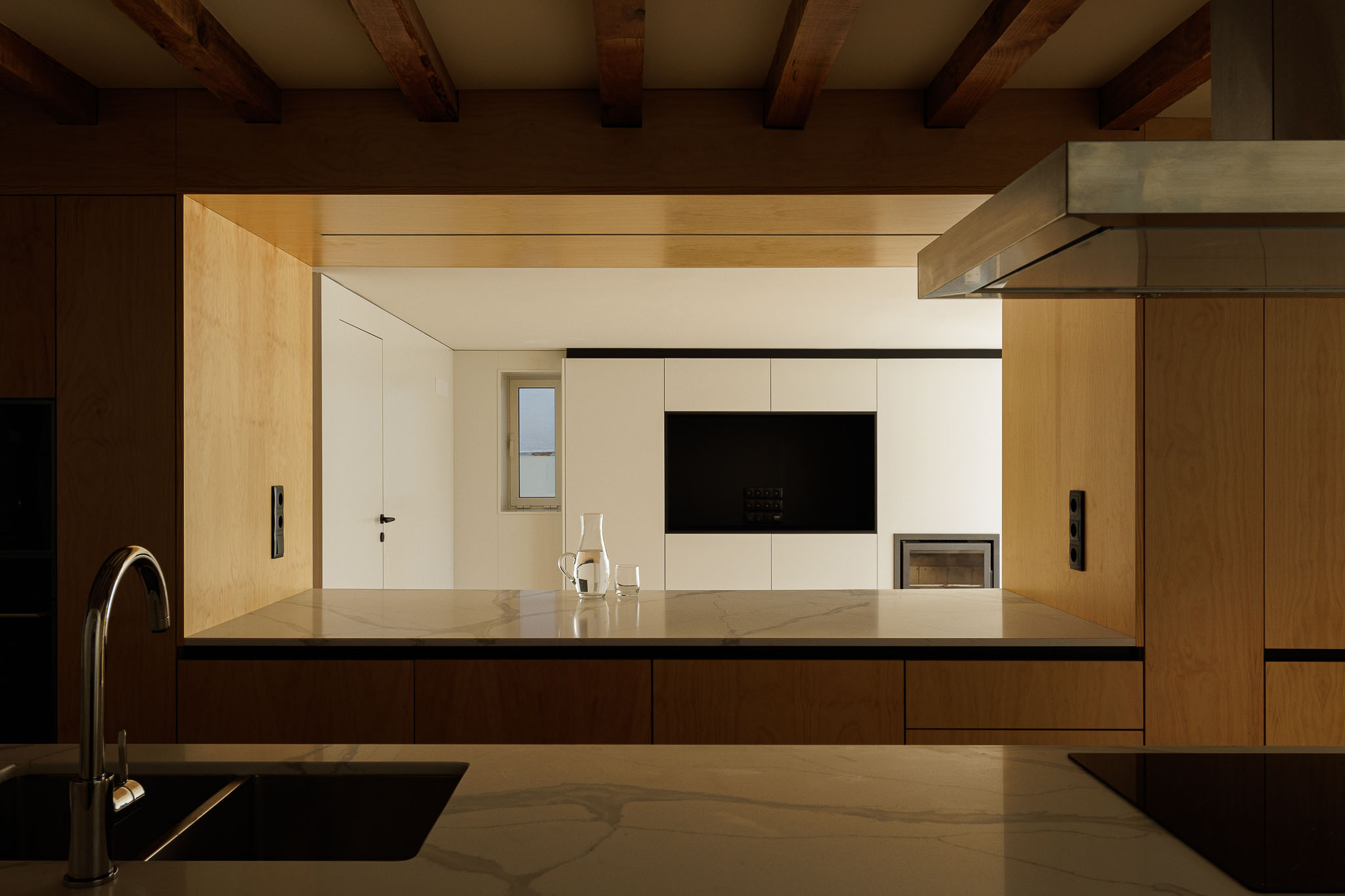 Cozinha: Vista da cozinha para a sala de estar com lareira e armários em madeira clara. — Casa dos anos 40 arquitetura Frari em Ílhavo