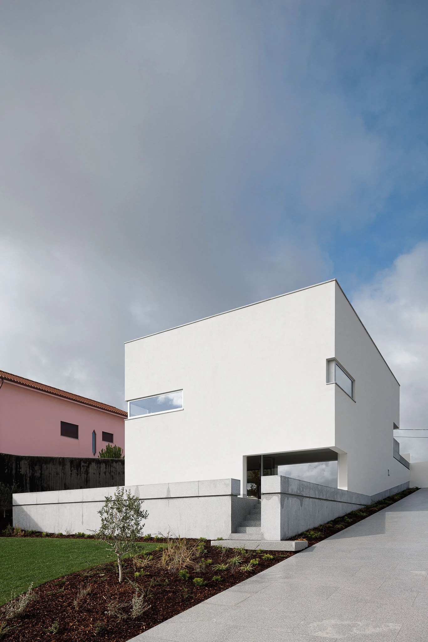 Vista exterior da casa cúbica branca com base em granito, jardim relvado e rampa de acesso em granito. — Casa em Santa Marinha arquitetura Helder da Rocha Arquitectos em Lousada