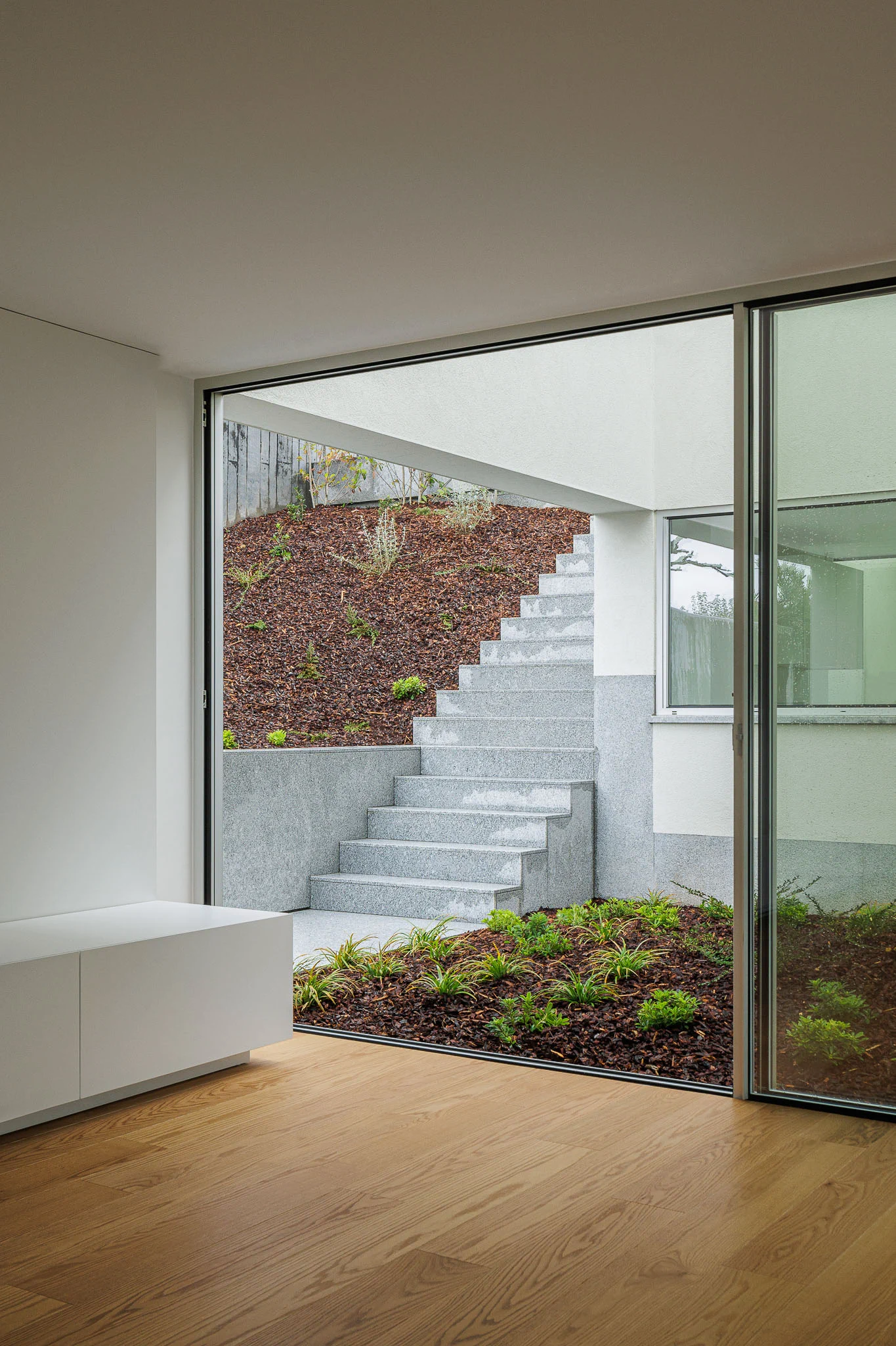 Interior com vista para escada exterior em granito, através de porta de correr envidraçada, com pavimento em madeira. — Casa em Santa Marinha arquitetura Helder da Rocha Arquitectos em Lousada